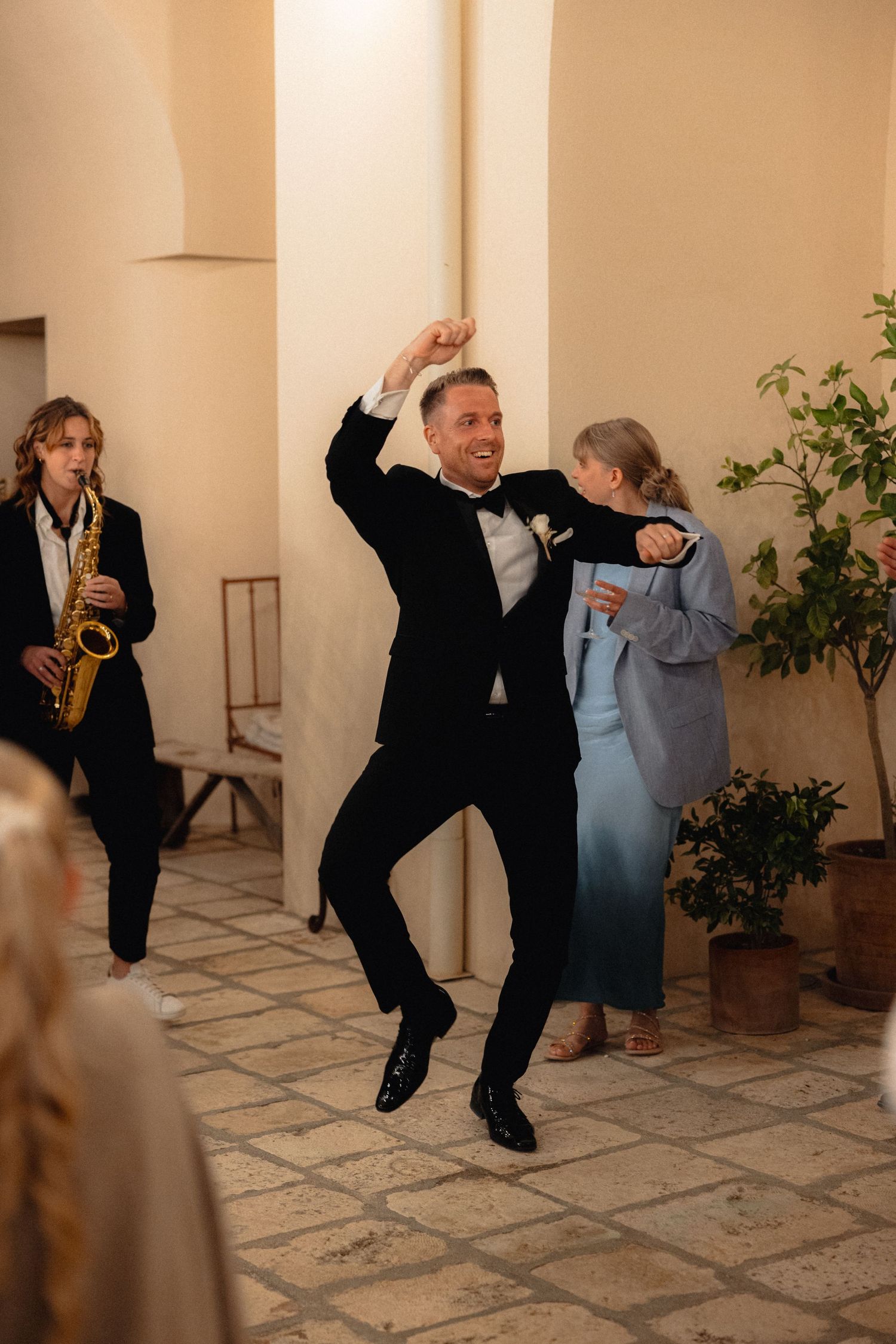 Musicians in black suits dance and perform in an elegant hallway with columns.