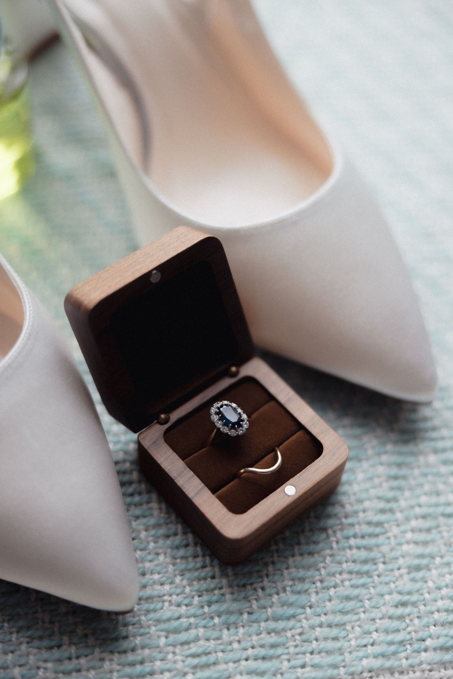 Diamond engagement ring in wooden box next to white bridal shoes on blue textile.