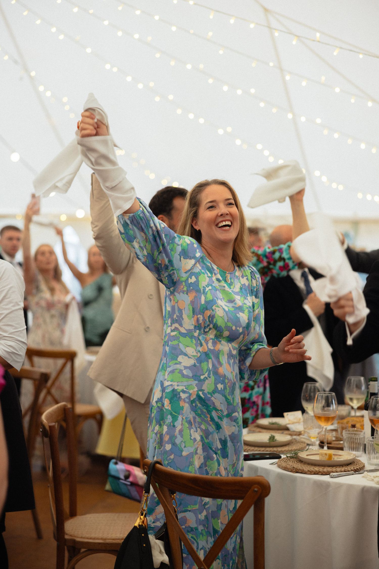 Guest in floral dress stands on chair celebrating at wedding reception with string lights overhead.