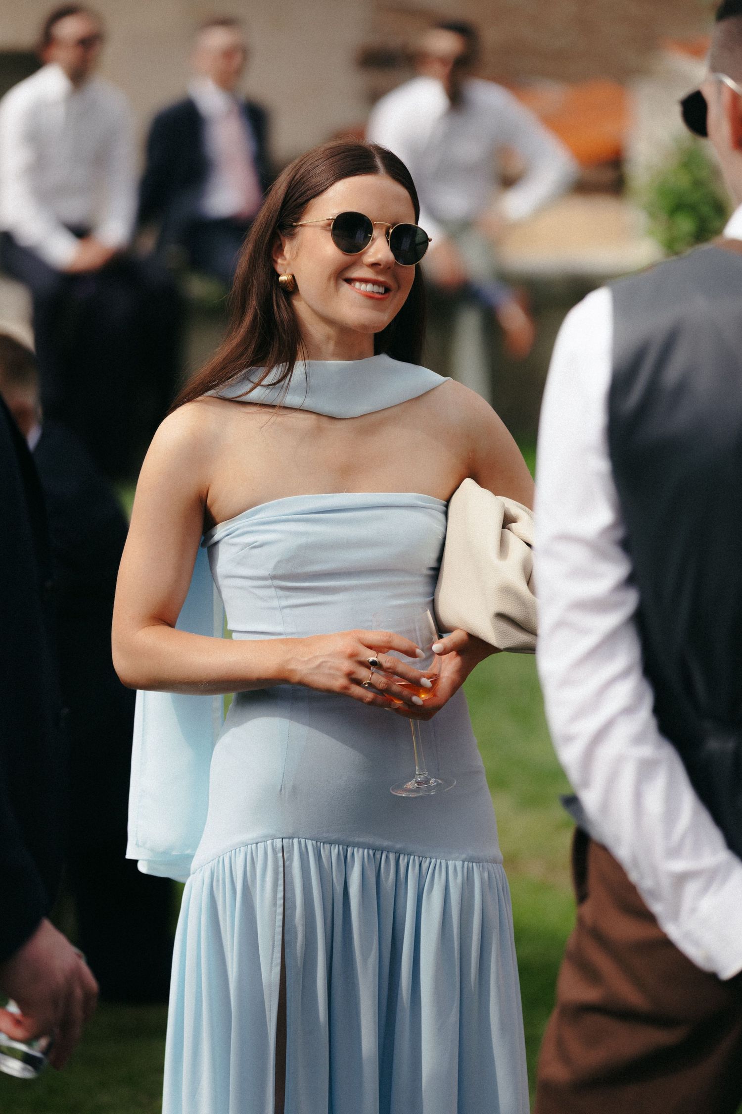 Guest in pale blue dress smiles while holding drink at outdoor reception.