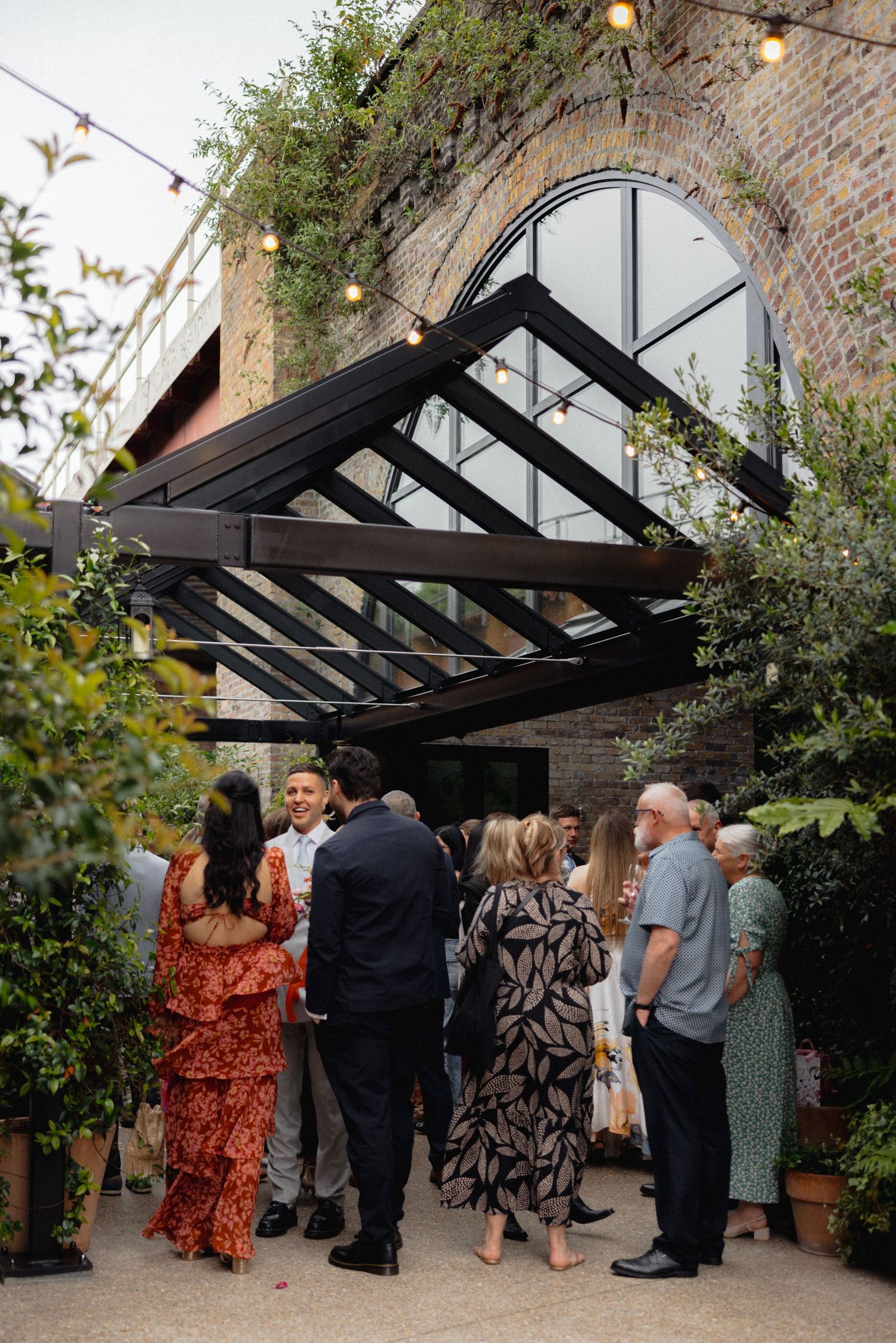 Wedding guests mingling under a glass-roofed courtyard with exposed brick walls and hanging lights.