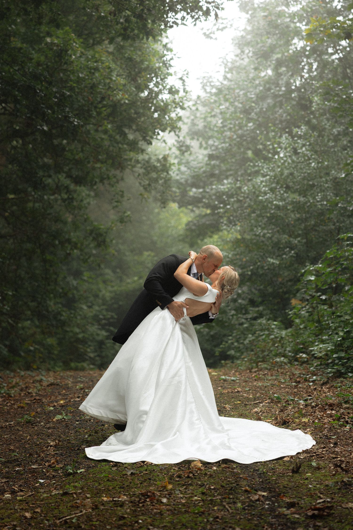 Couple shares intimate dance pose in misty woodland with flowing white wedding gown.