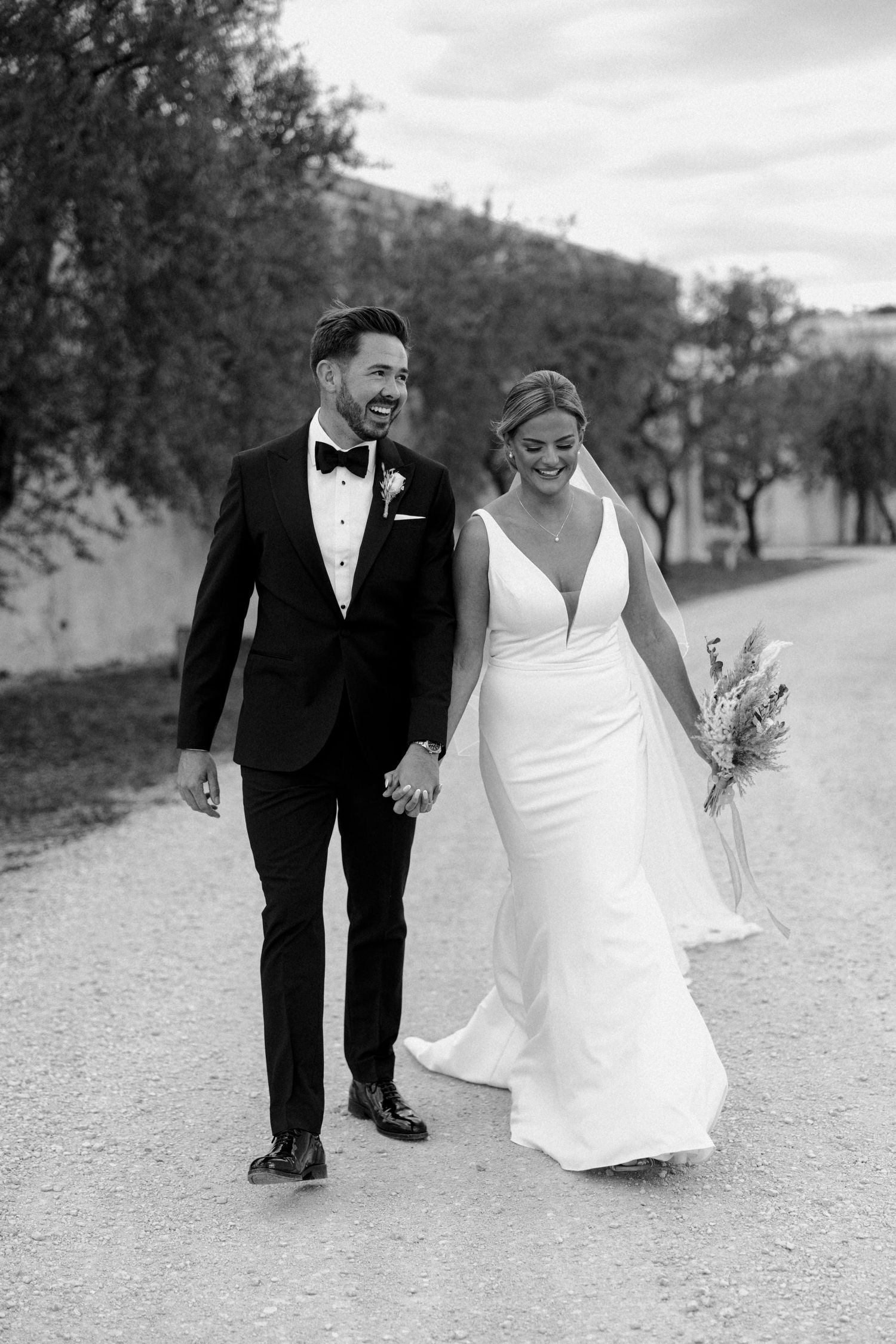 A wedding couple walks together on a gravel path in black and white, with the bride's dress trailing behind.
