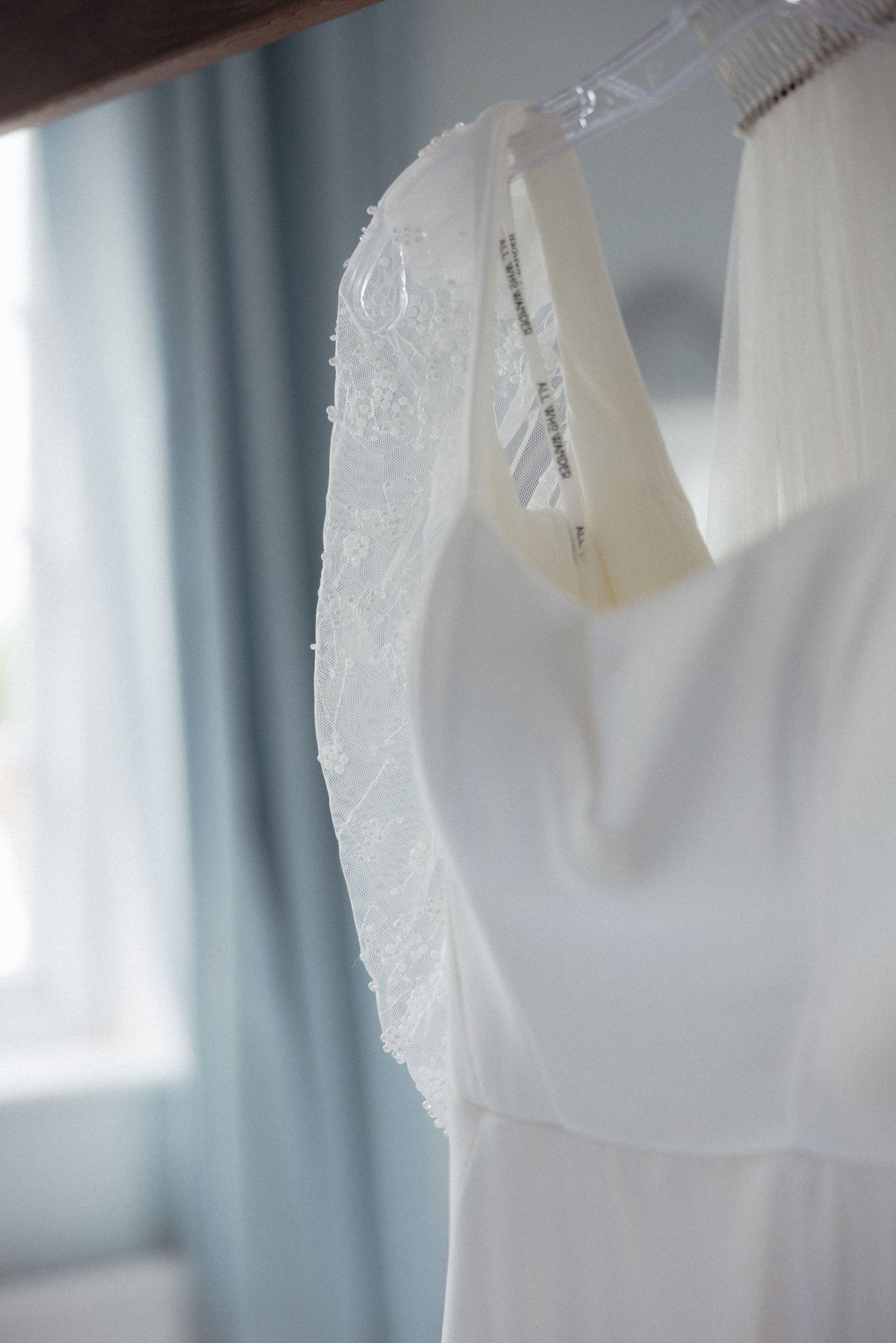 White lace wedding dress detail hanging against soft blue curtains in natural light.