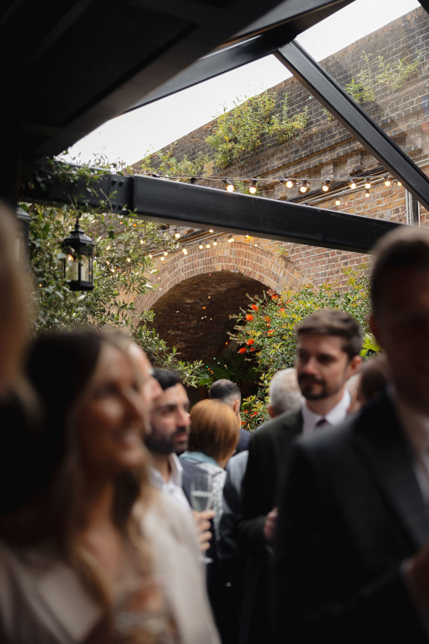 Wedding guests gather under a brick archway with string lights at an outdoor venue.