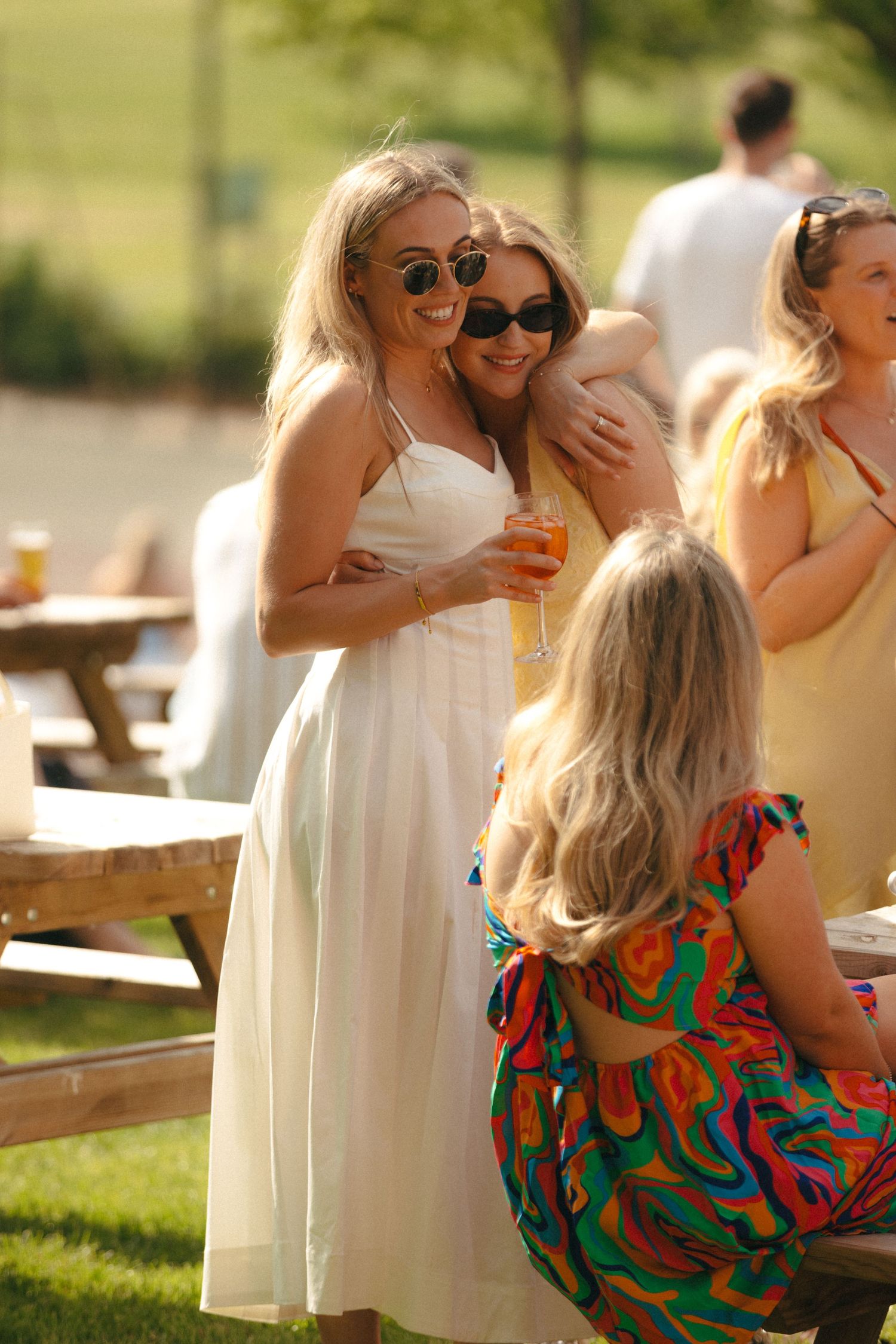 Guests enjoying drinks and conversation at an outdoor summer party with wooden picnic tables.