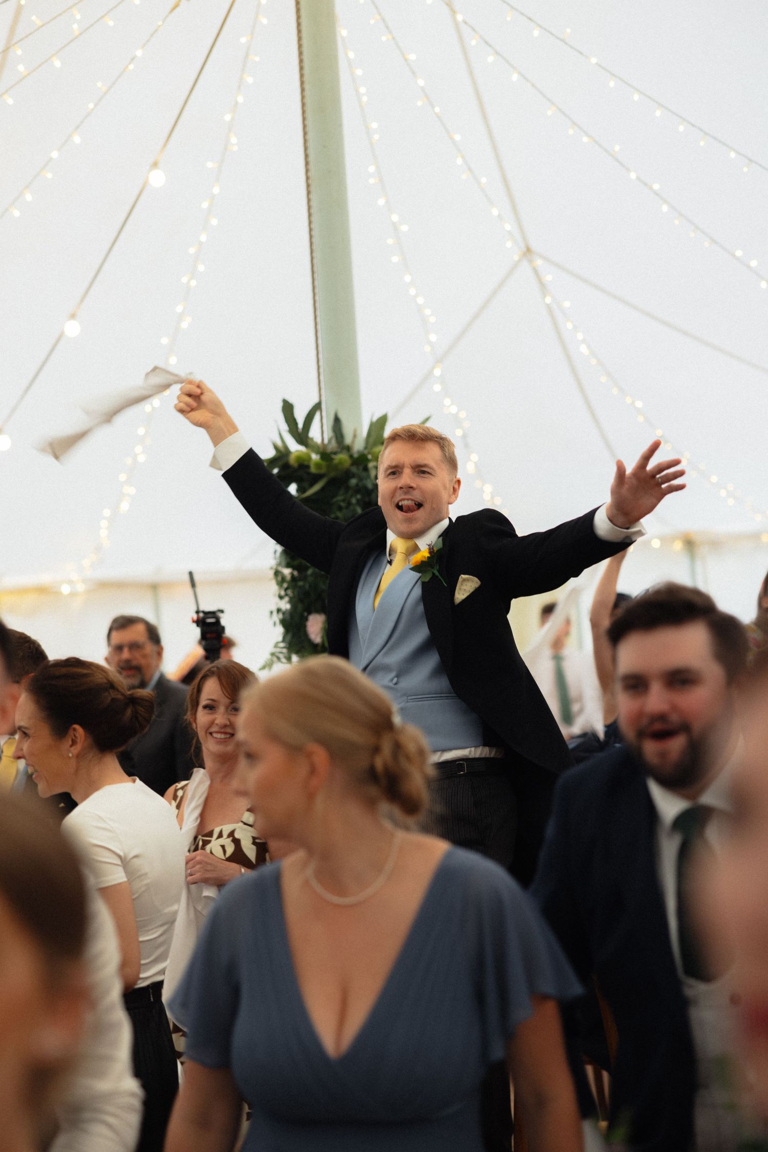 Wedding guest stands on chair celebrating during reception under tent with string lights.