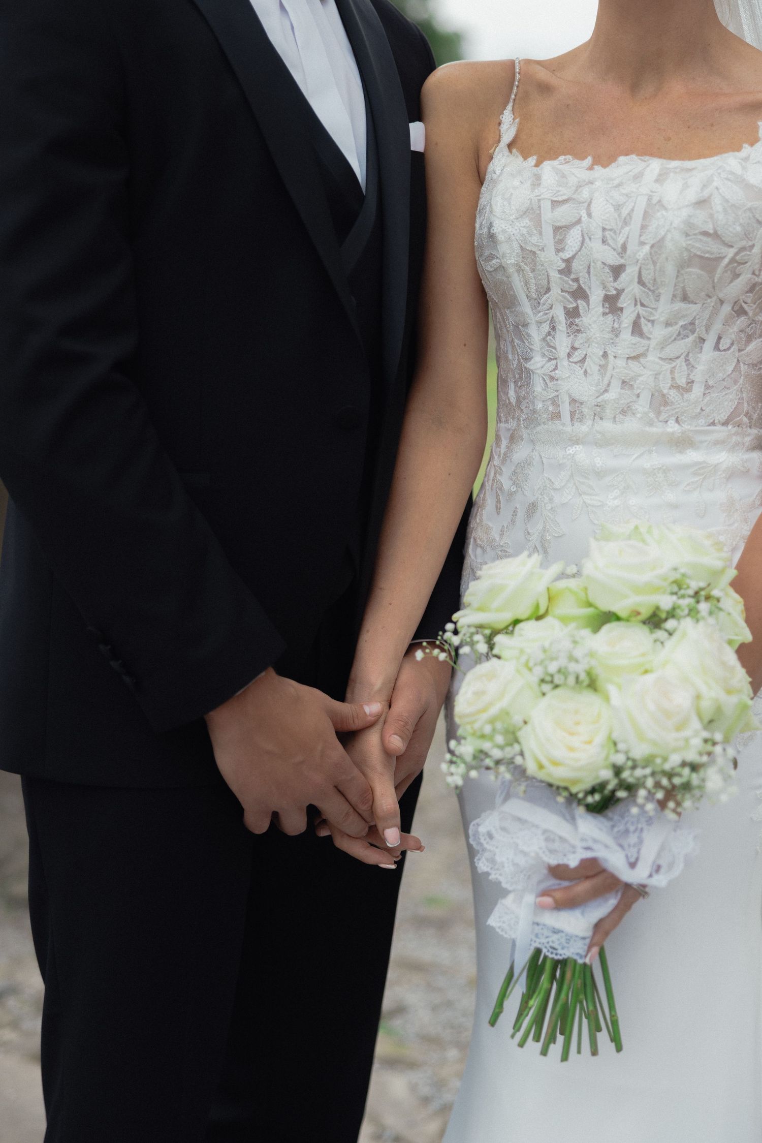 Close-up of couple holding hands with white rose bouquet and detailed lace wedding dress.