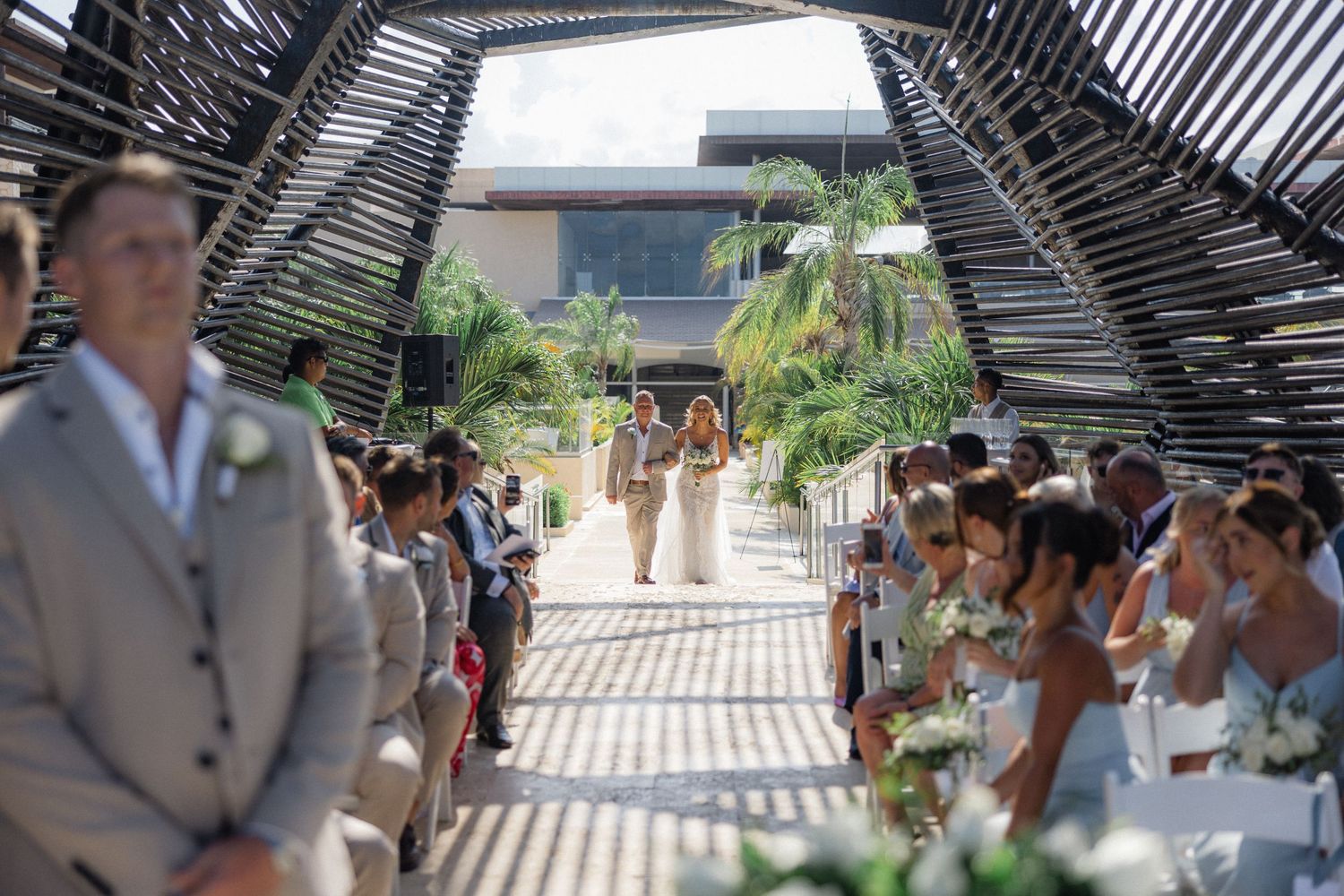Guests gather under a modern pergola structure during an outdoor wedding ceremony.