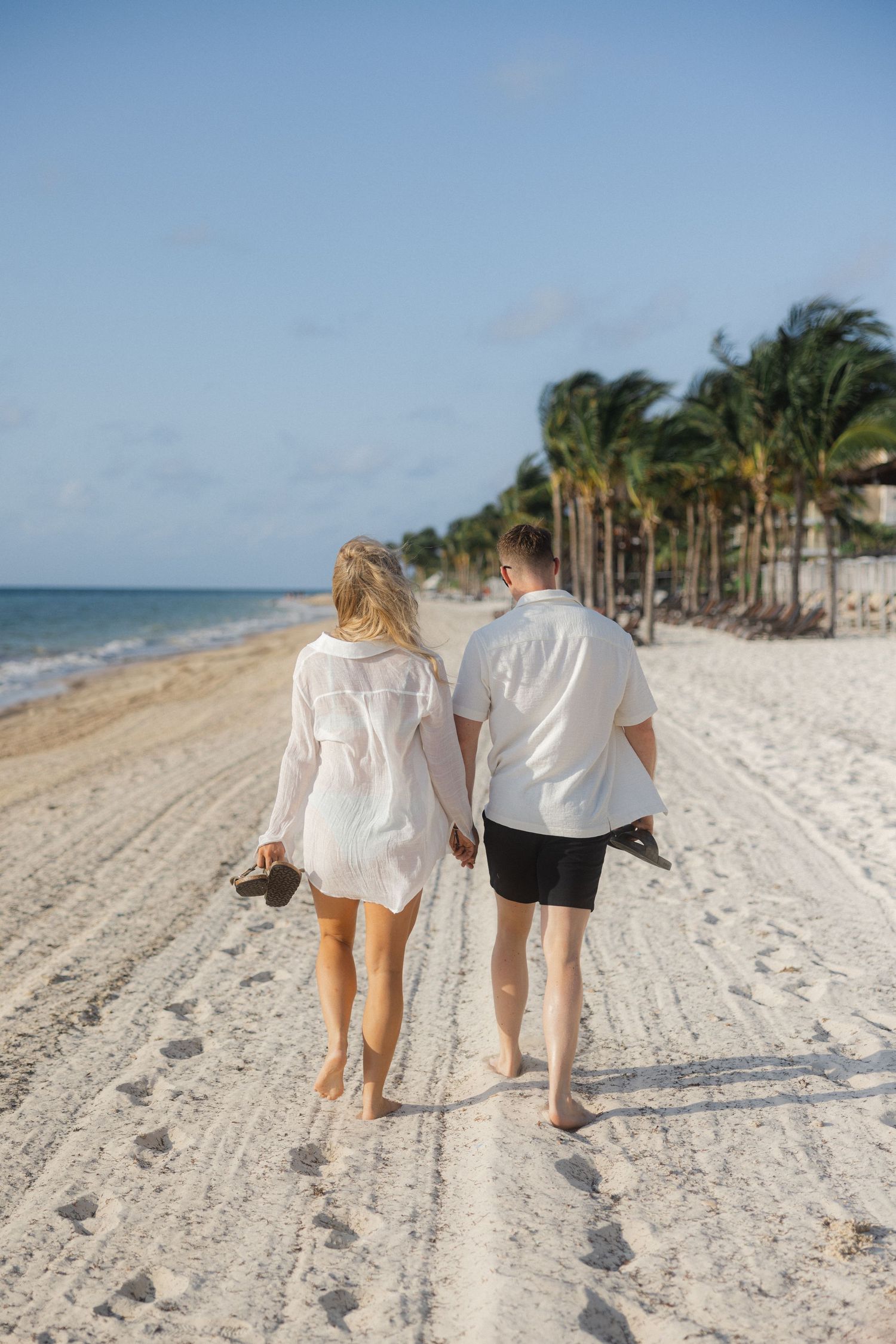 Two people walking hand in hand along a pristine white sand beach lined with palm trees.