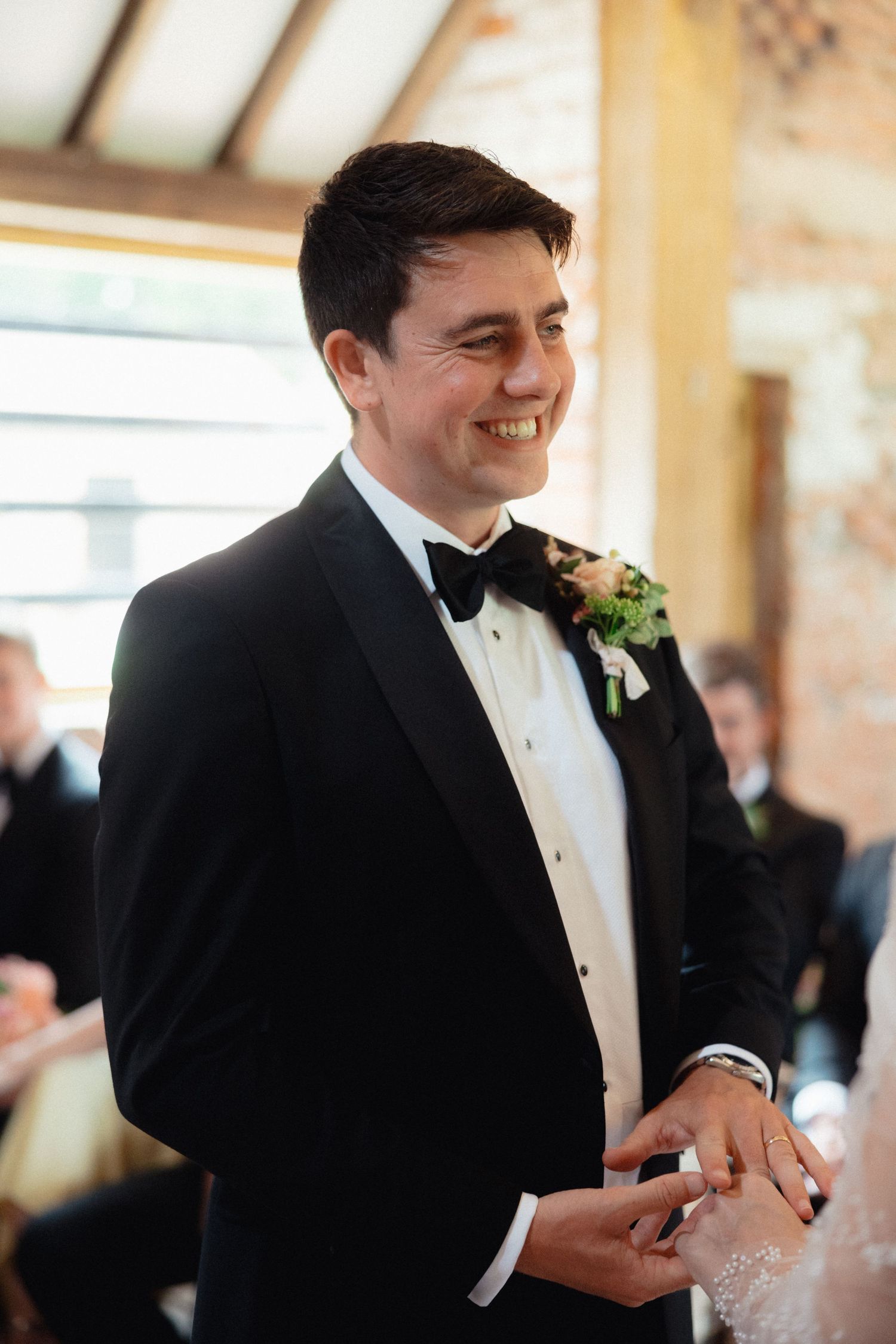Groom in black tuxedo smiling during wedding ceremony in elegant indoor setting.