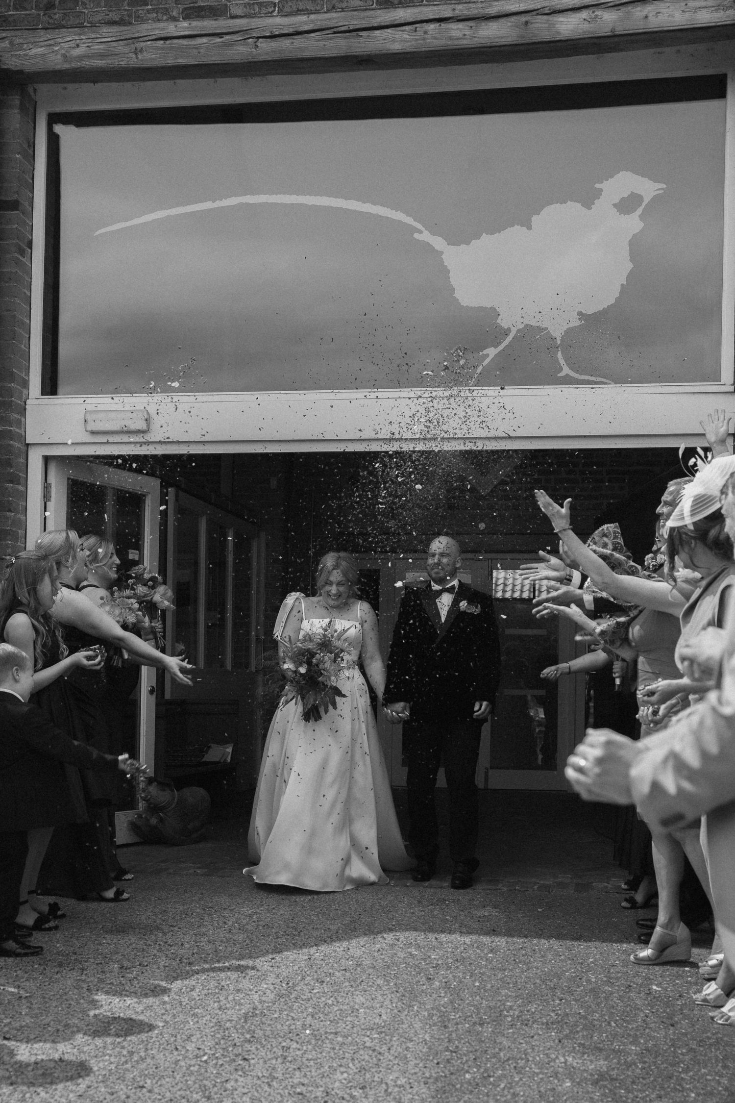 Black and white photo of newlyweds exiting venue while guests throw confetti.