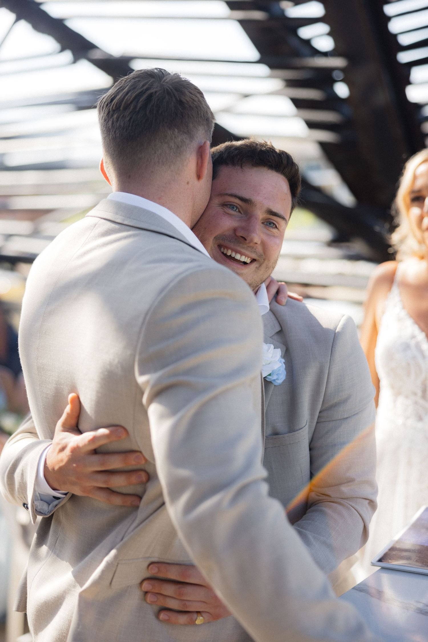 Groomsmen in light suits share emotional embrace during wedding celebration.