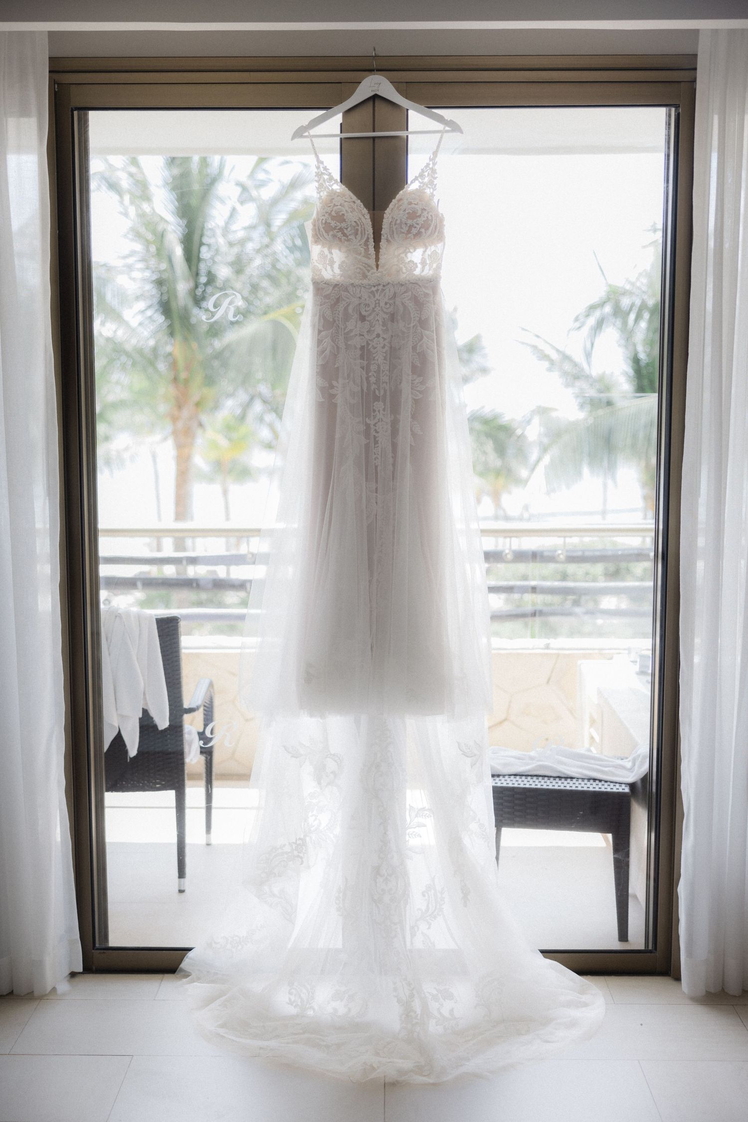 White wedding dresses hanging in front of large windows with tropical palm trees visible outside.