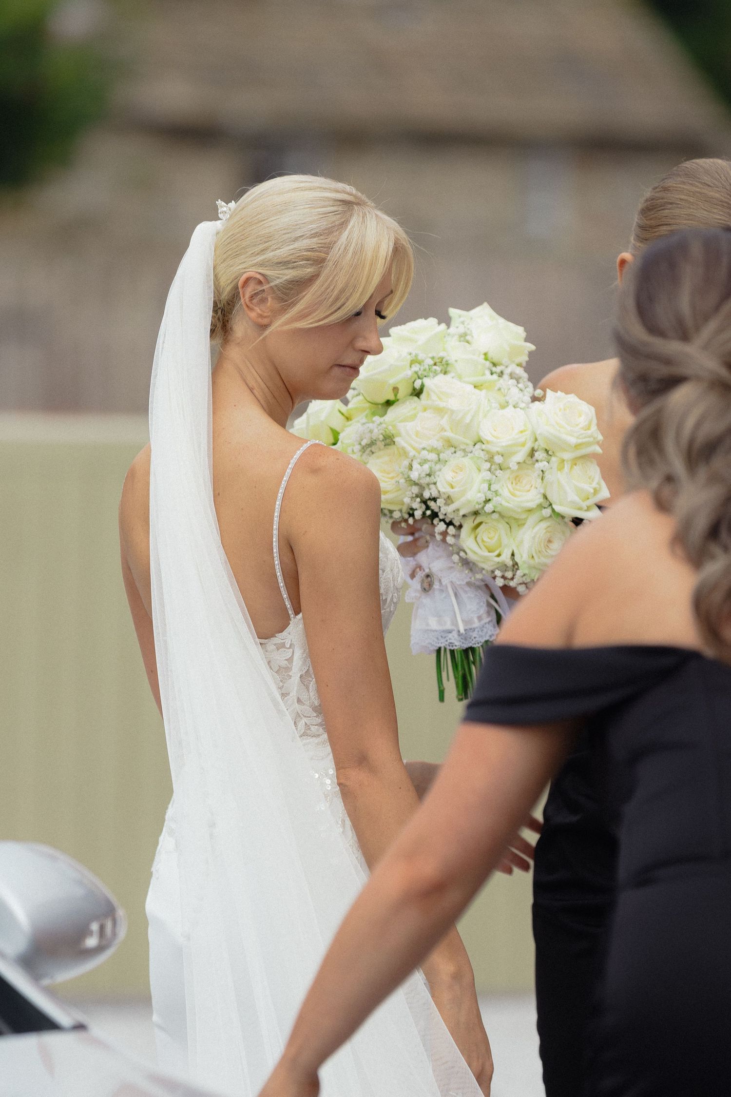 A bride in a flowing white gown carries a bouquet of white flowers during an outdoor ceremony.