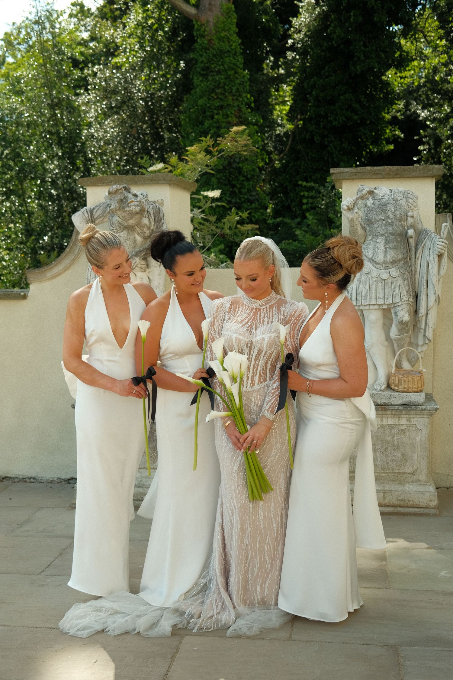 Four women in elegant white dresses stand together at an outdoor wedding venue with decorative stone pillars.