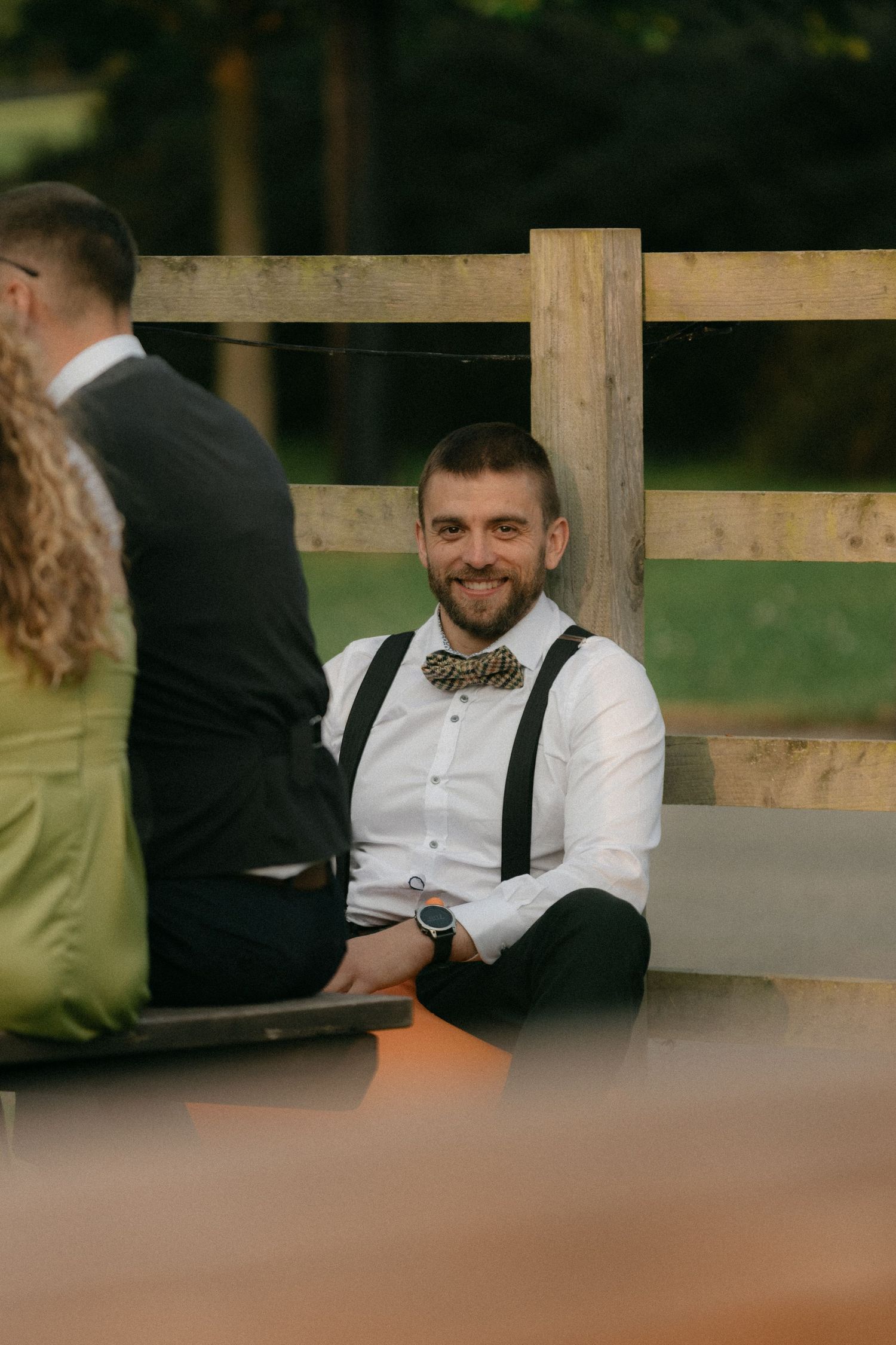 A guest in suspenders and bow tie relaxes at an outdoor wedding reception.