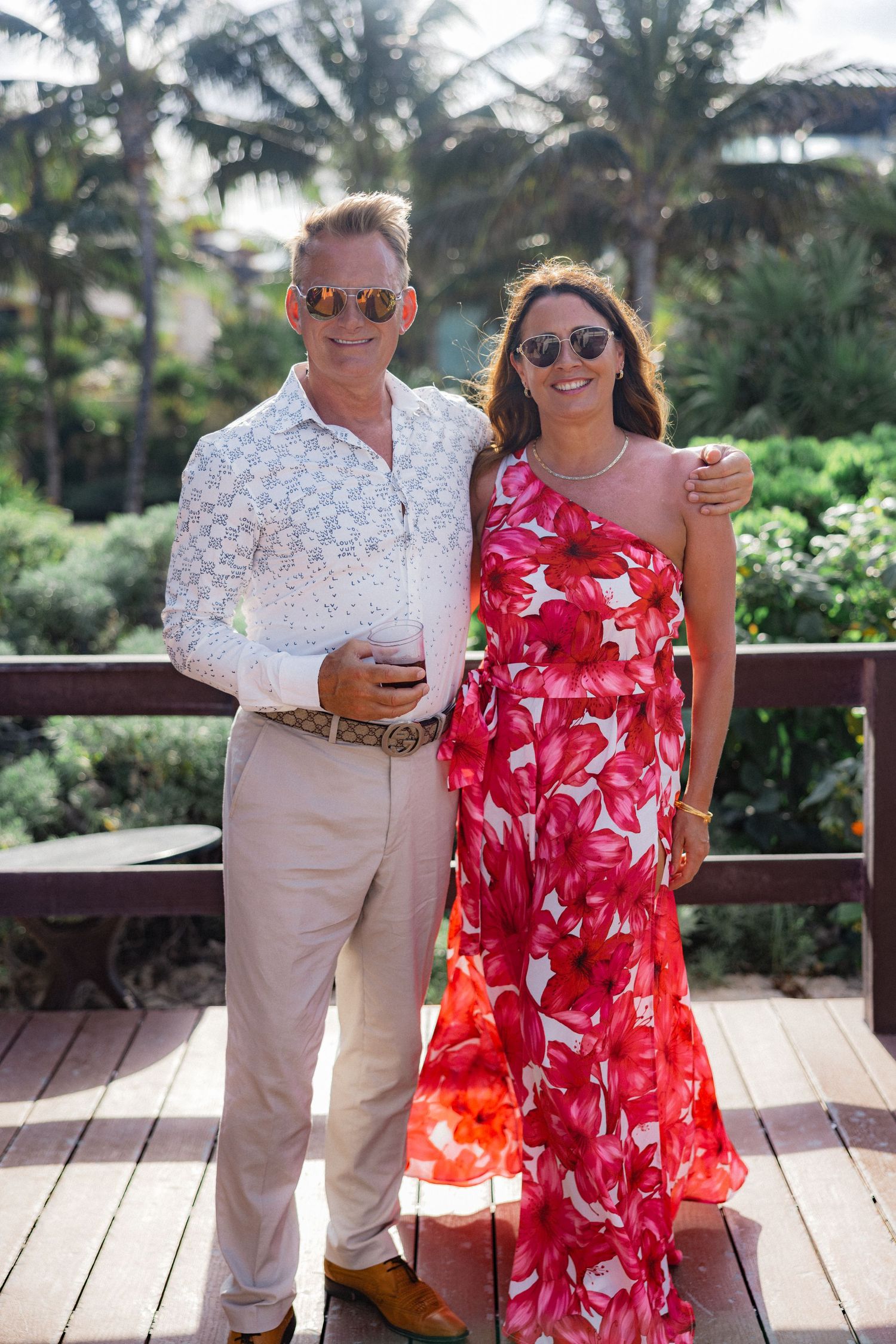 Guests in tropical attire enjoy drinks on wooden deck with palm trees in background.