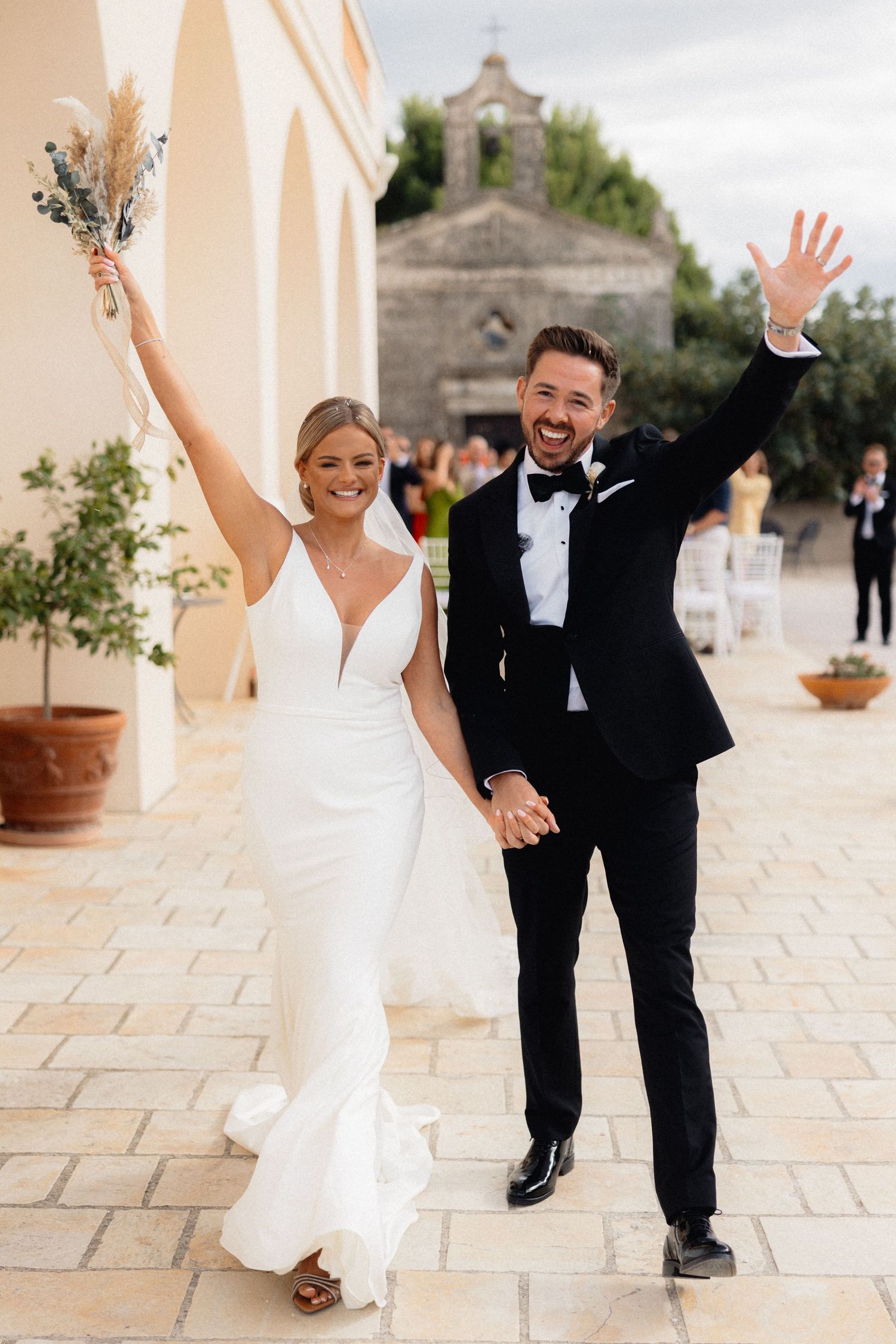 Newlyweds celebrating with raised arms on a sunny stone terrace.