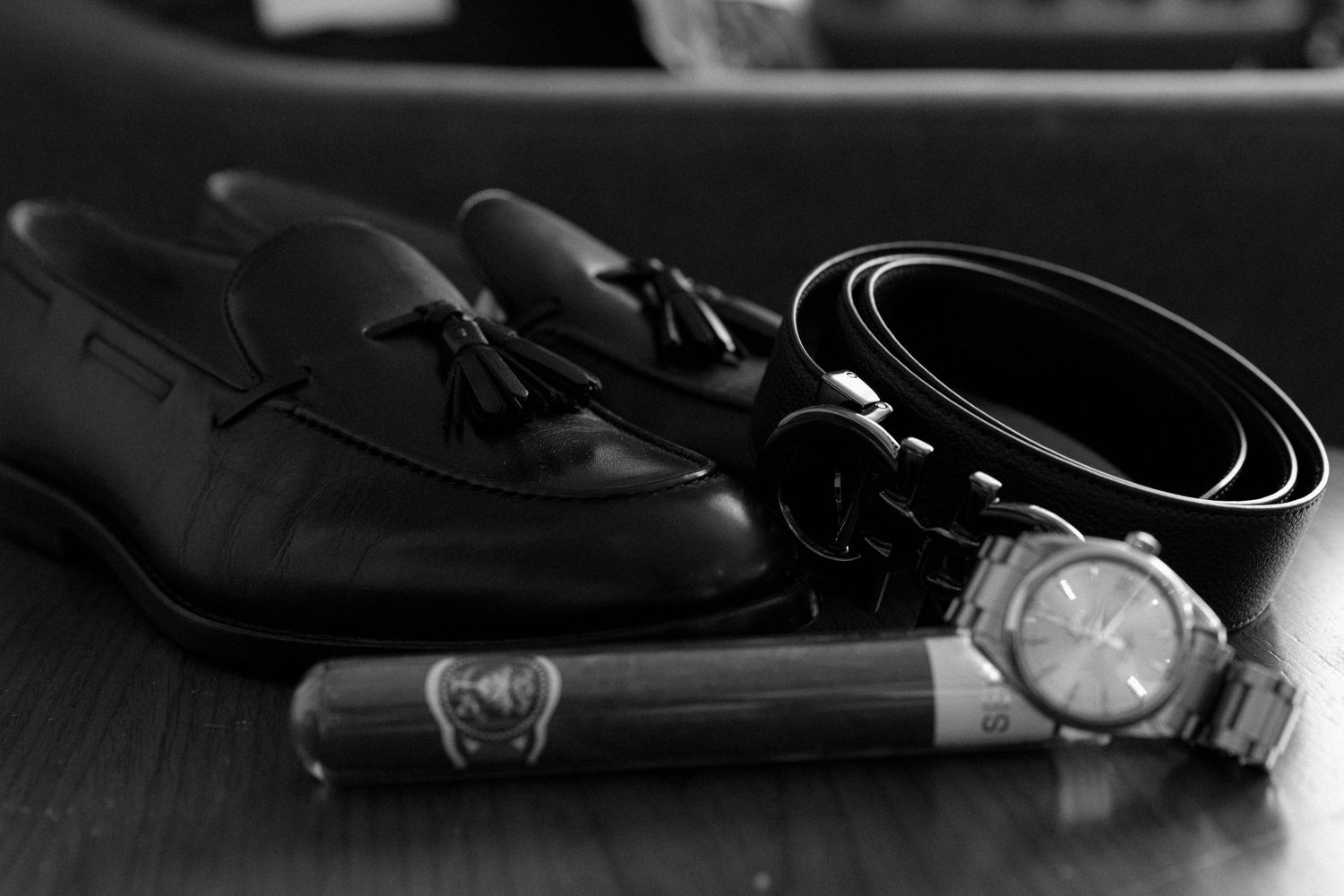 Black dress shoes, watch, belt and cigar arranged on dark wooden surface in monochrome style.
