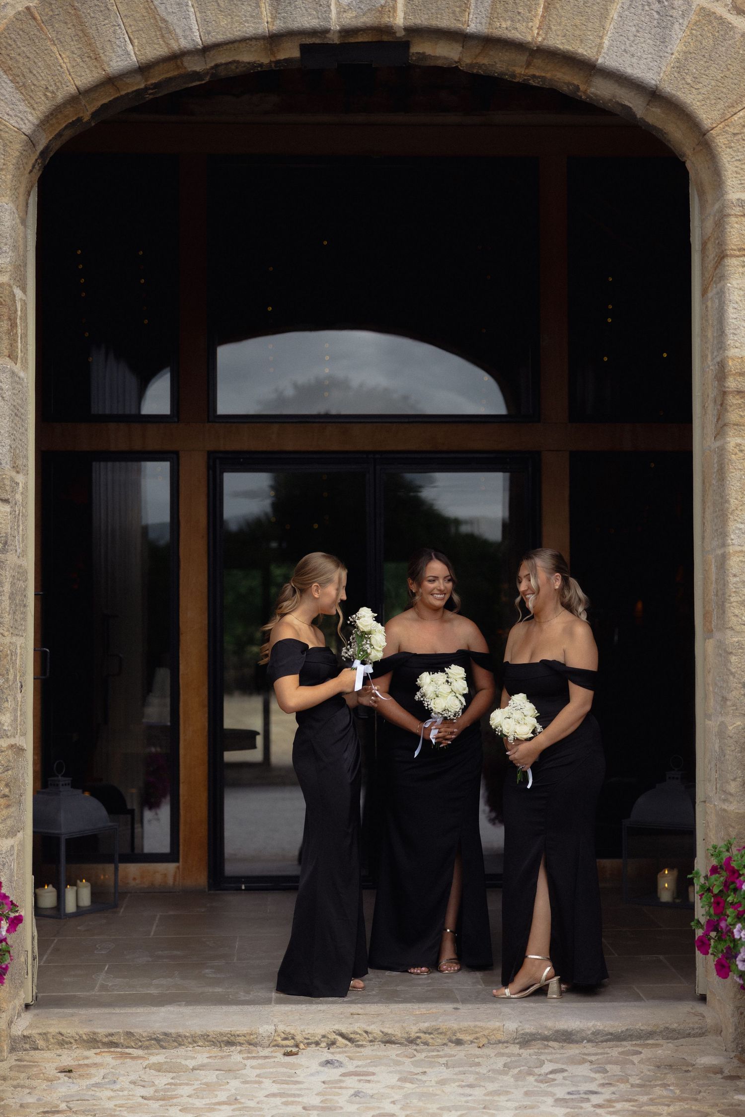 Bridesmaids in black dresses gather under stone archways at an elegant venue.