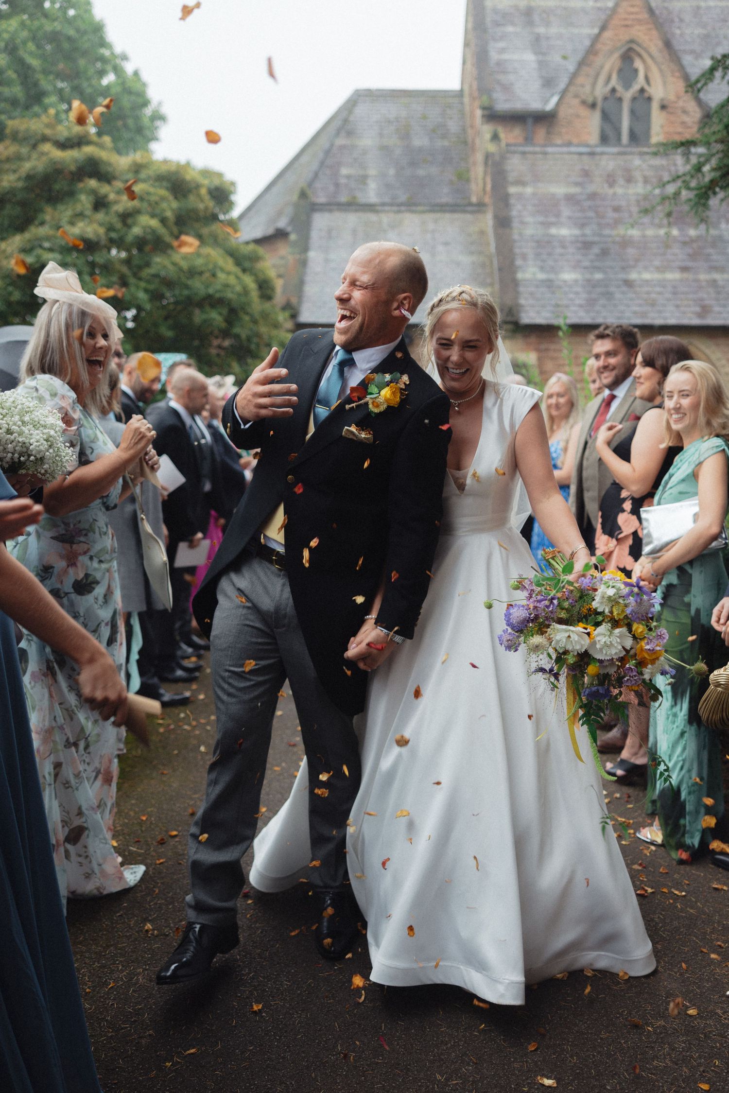 Wedding party celebrates with colorful confetti toss outside historic stone church building.