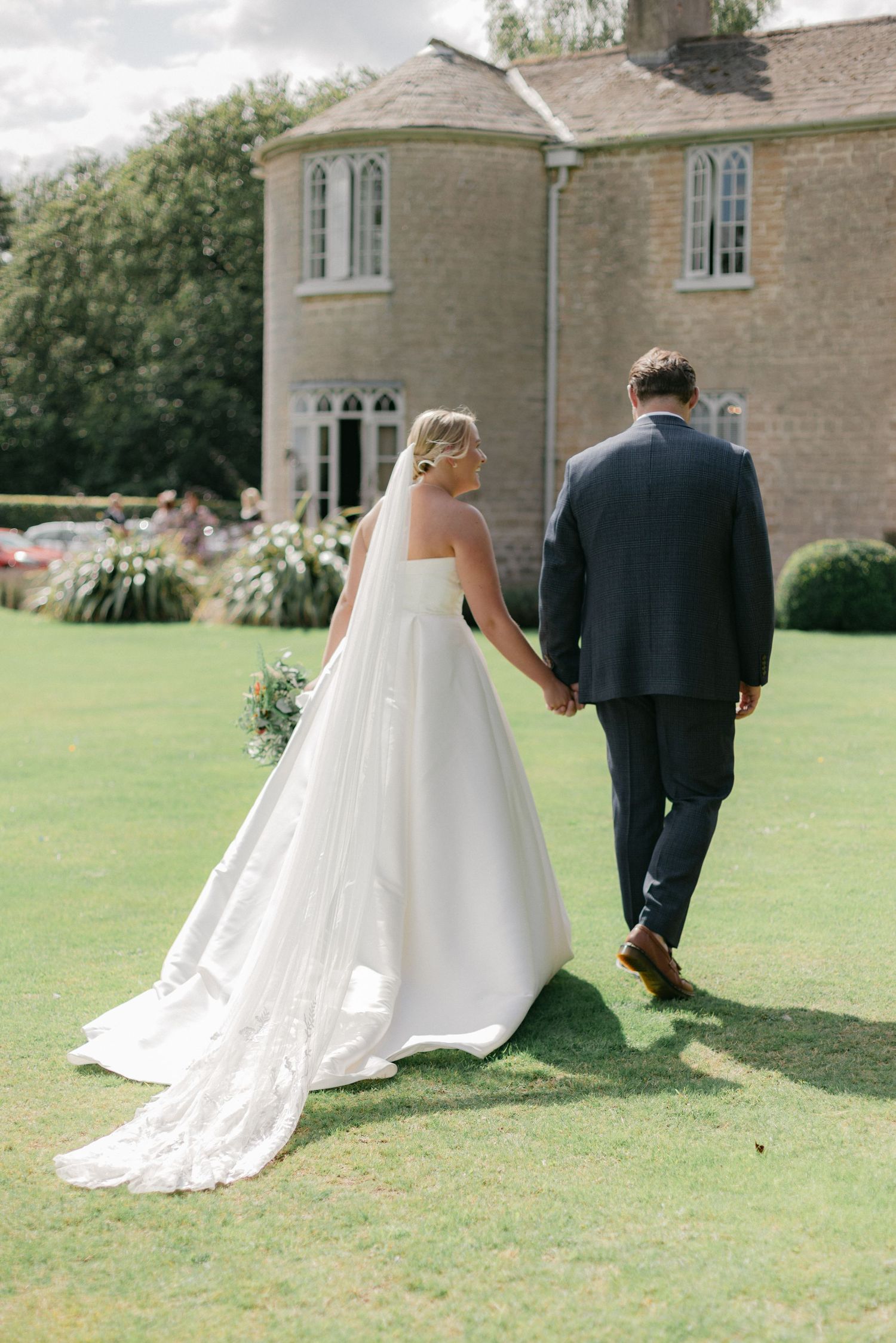 A wedding couple photographed from behind walking across a manicured lawn at a stately venue.