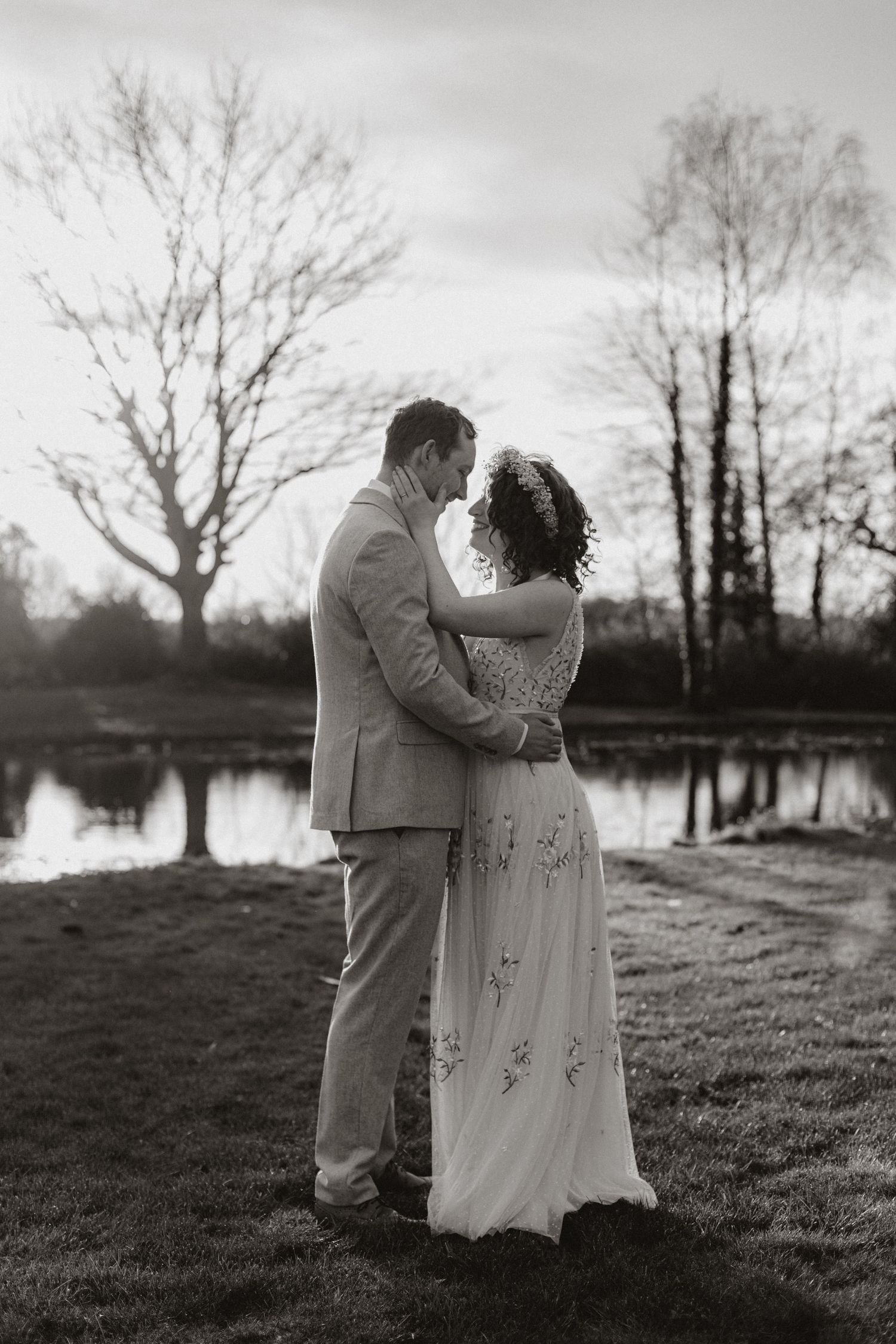 Black and white portrait of newlyweds embracing by a lake with bare trees in background.