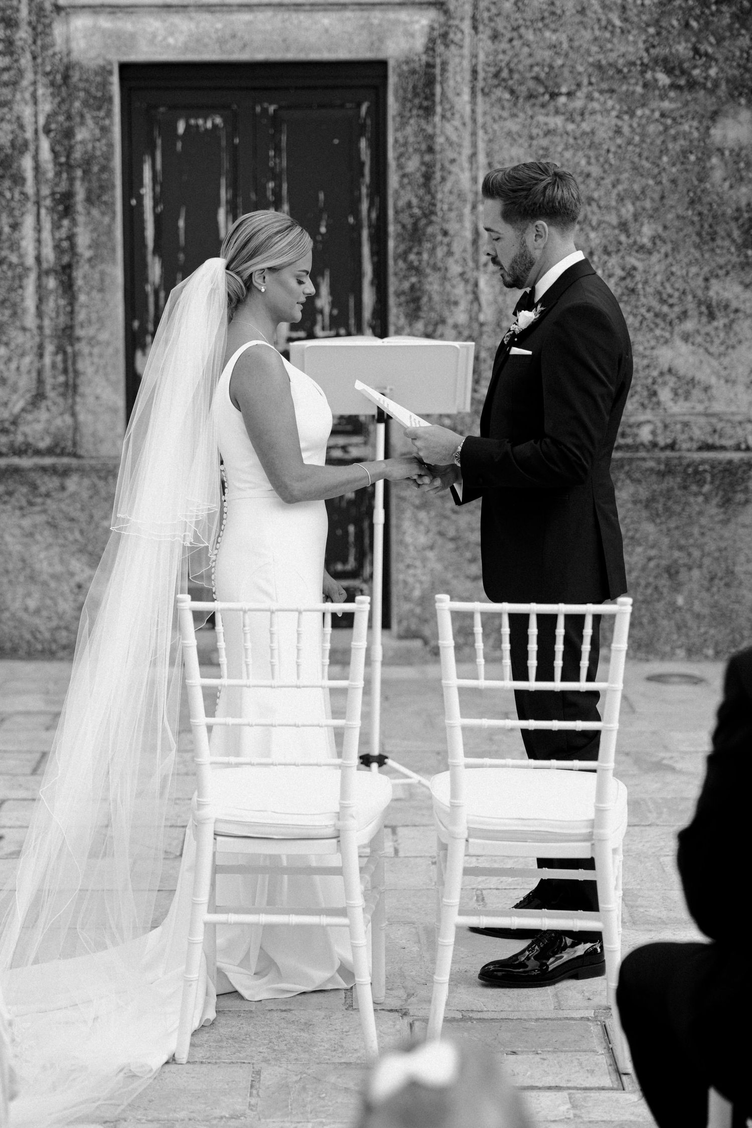 A wedding couple shares a playful moment between white chairs against a stone wall backdrop.