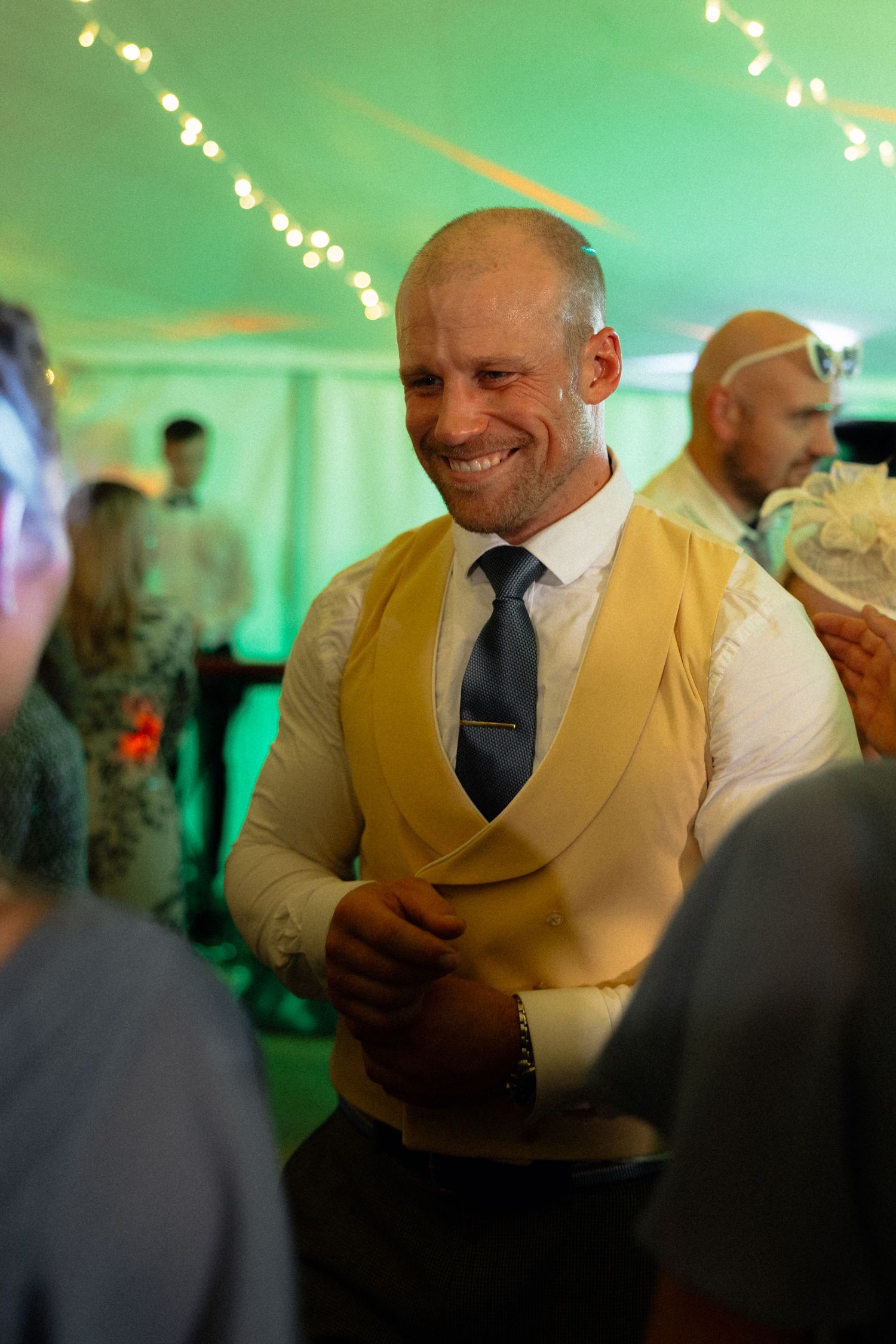 Wedding guest in gold vest and tie smiles during evening reception with green ambient lighting.