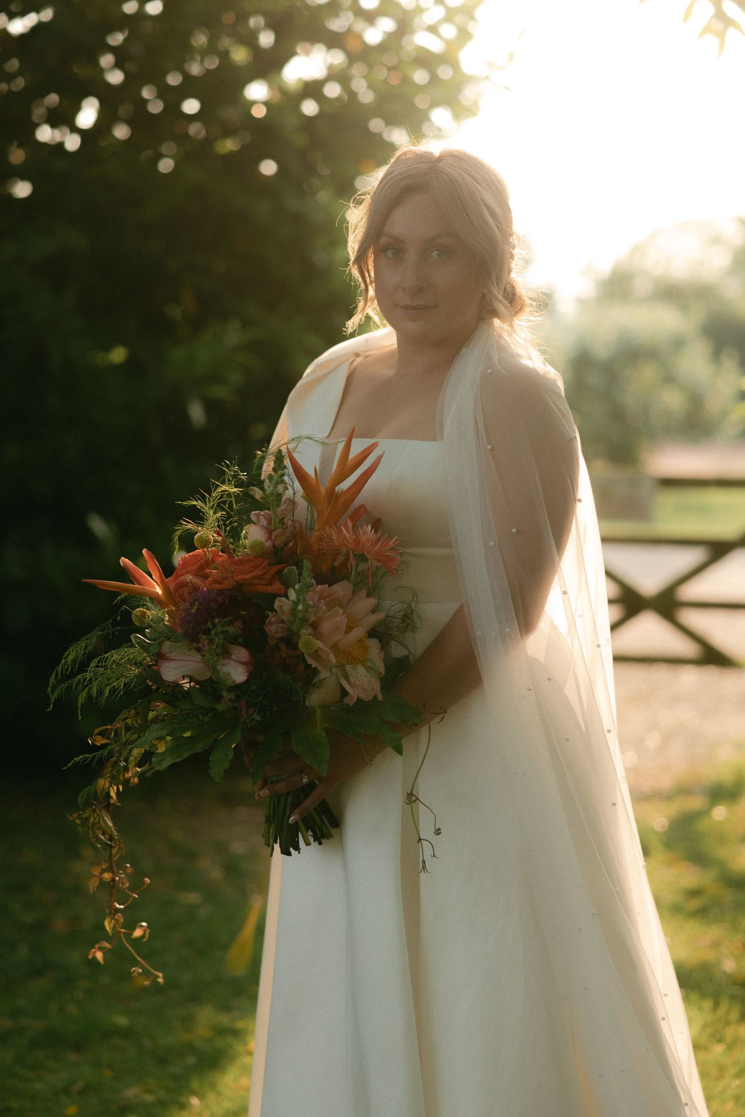 A bride in a white dress holds a vibrant orange and pink floral bouquet in golden sunlight.