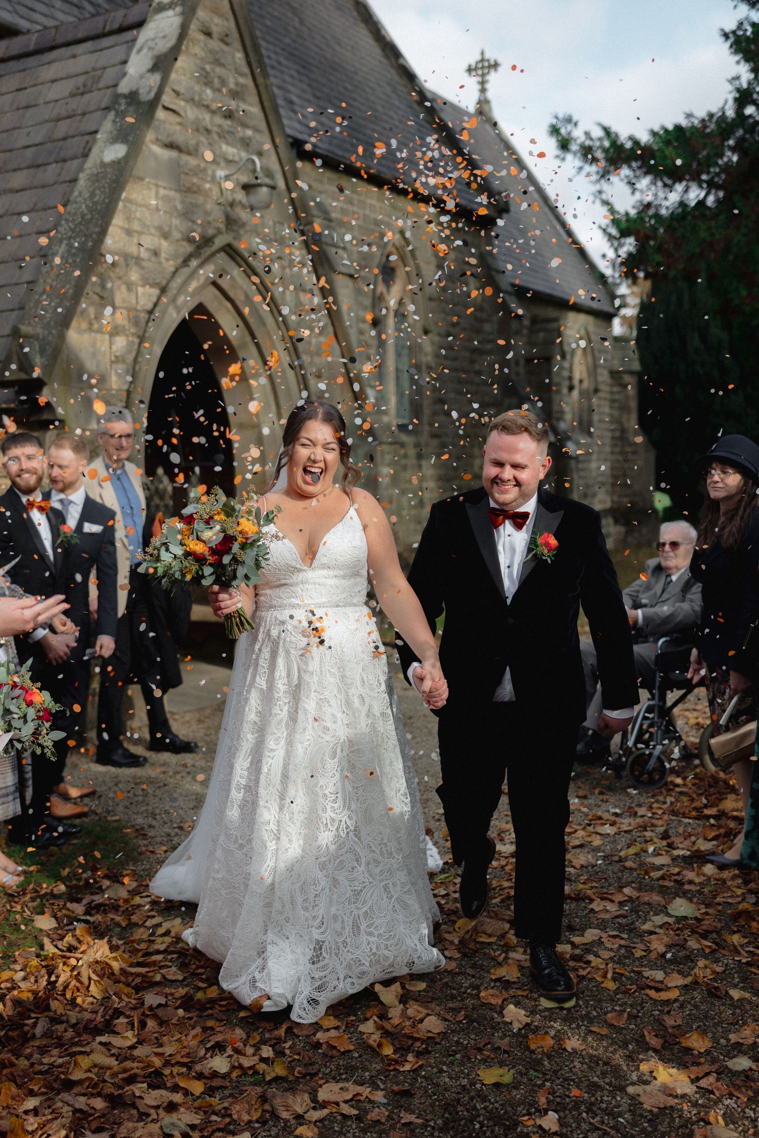 Joyful wedding exit with confetti outside stone church in autumn.