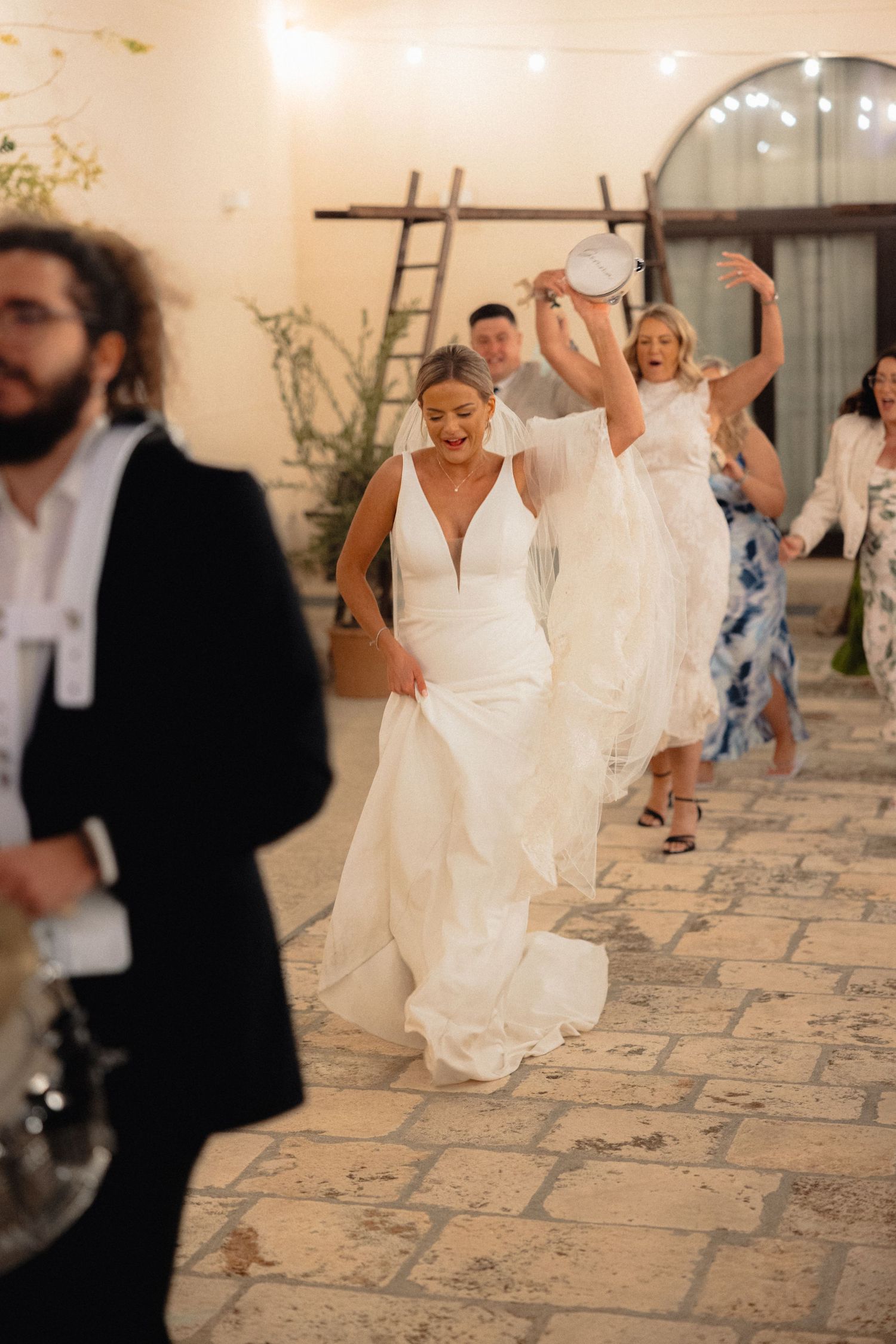 Wedding guests in formal attire dance and celebrate on a stone floor at an indoor reception.