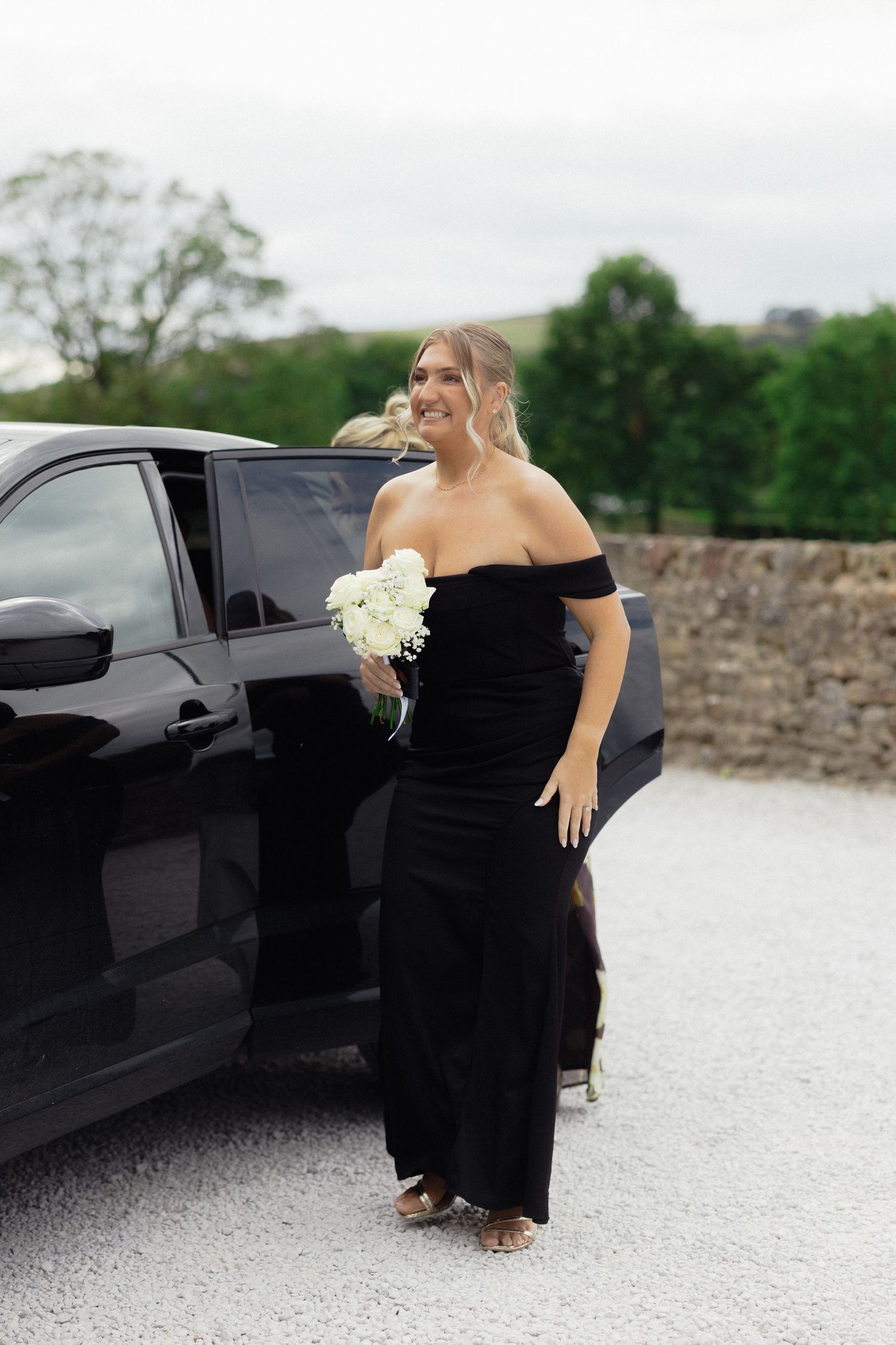 Wedding attendant in black off-shoulder dress holding white bouquet next to car.