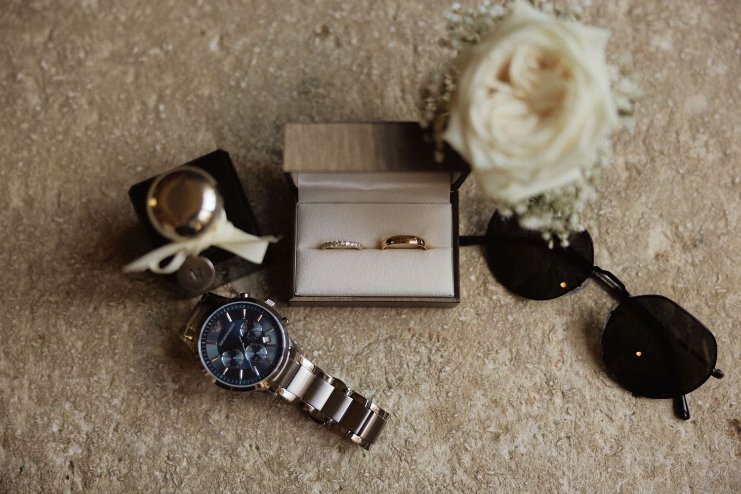 Wedding accessories including rings, watch and sunglasses arranged on textured surface.