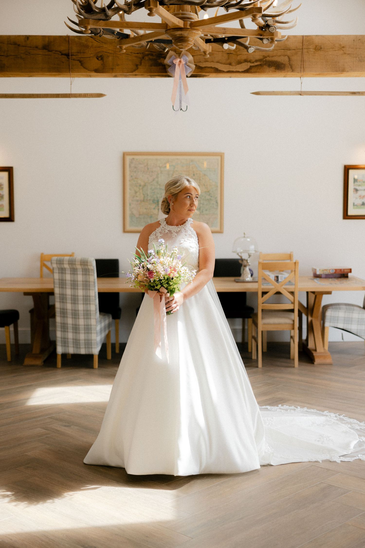 A bride in a flowing white gown stands in sunlit room with wooden beams and light walls.