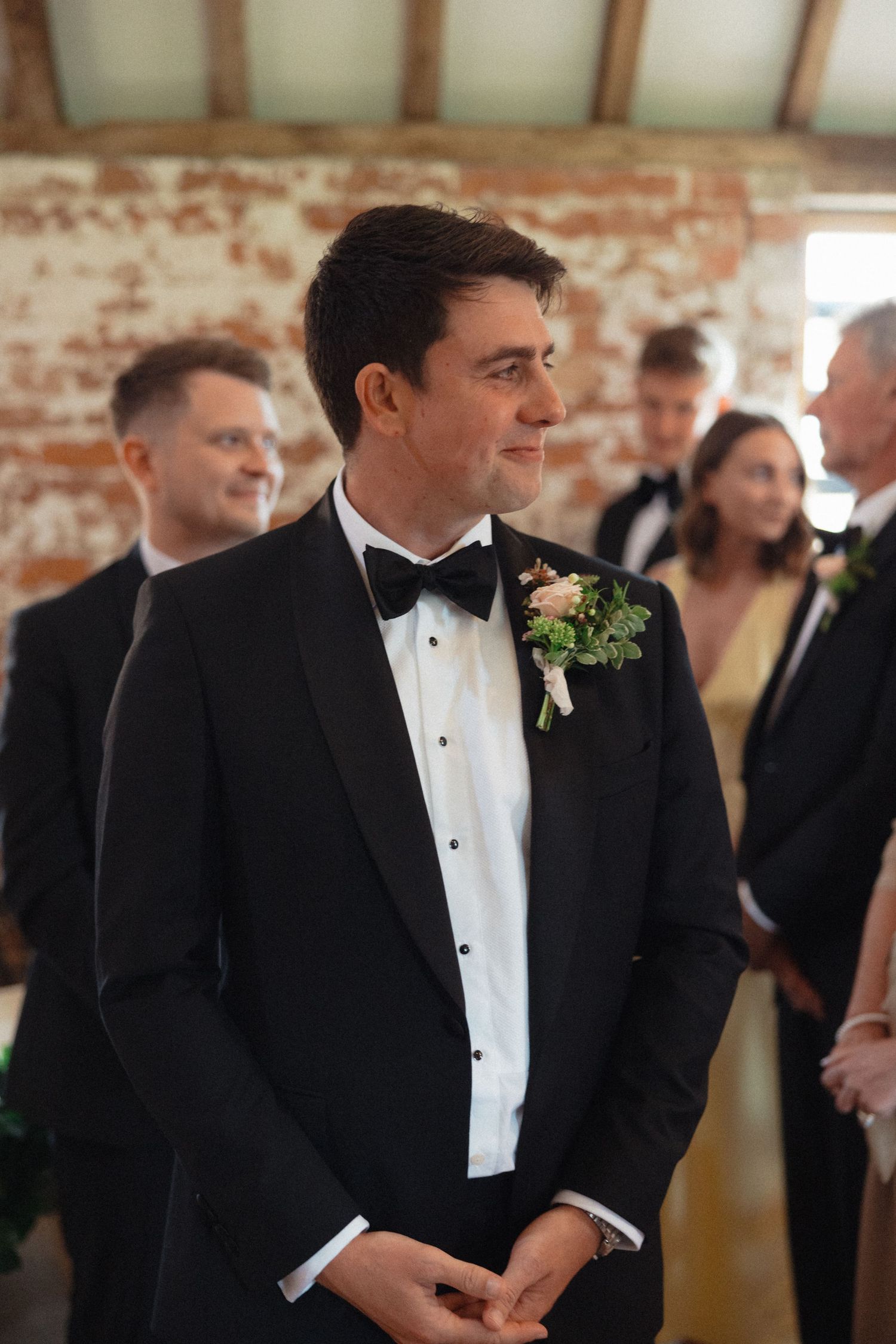 Groomsmen in tuxedos and boutonnieres stand in a rustic brick-walled venue.