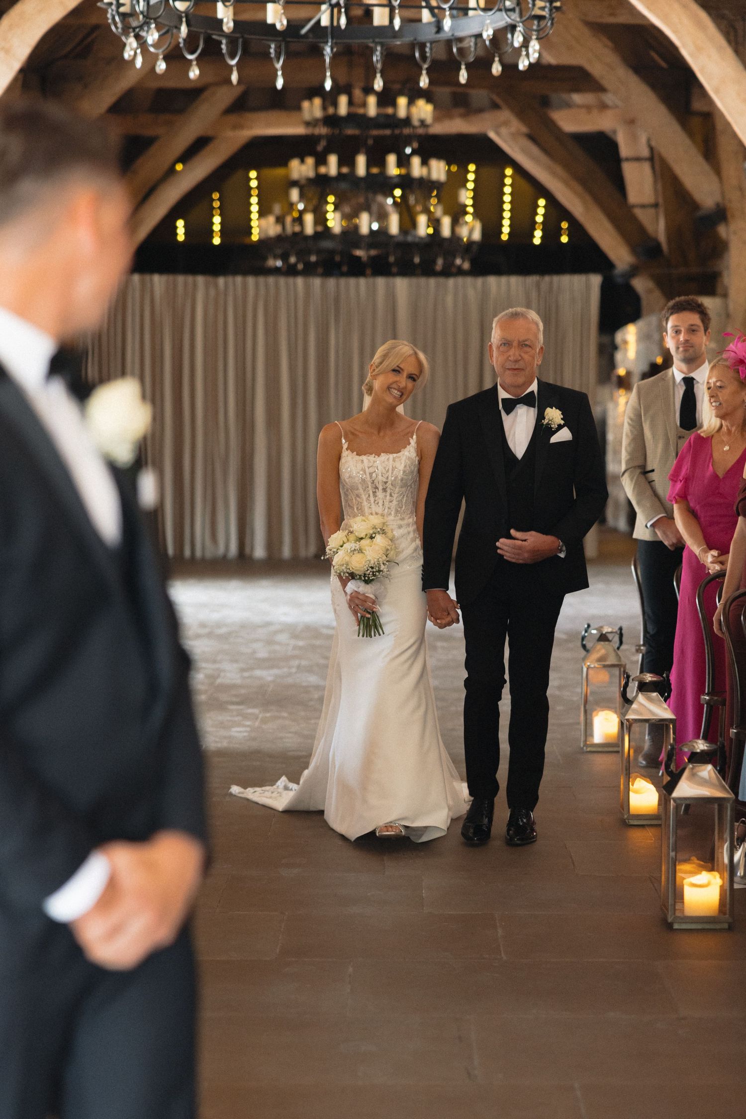 Wedding processional in rustic barn venue with lantern-lit aisle.