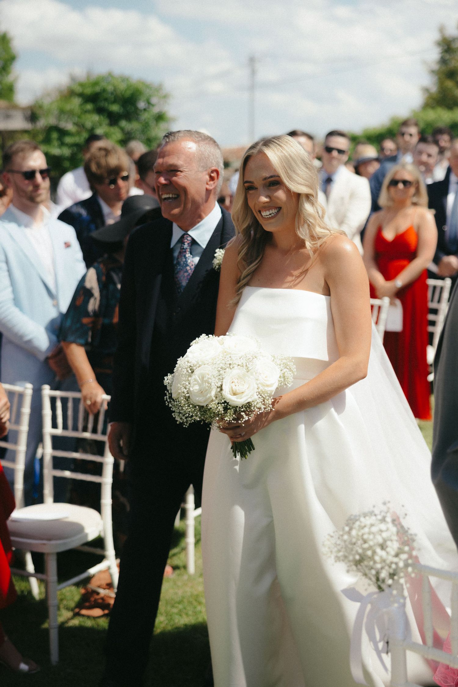 Joyful outdoor wedding ceremony with guests watching bride holding white bouquet.