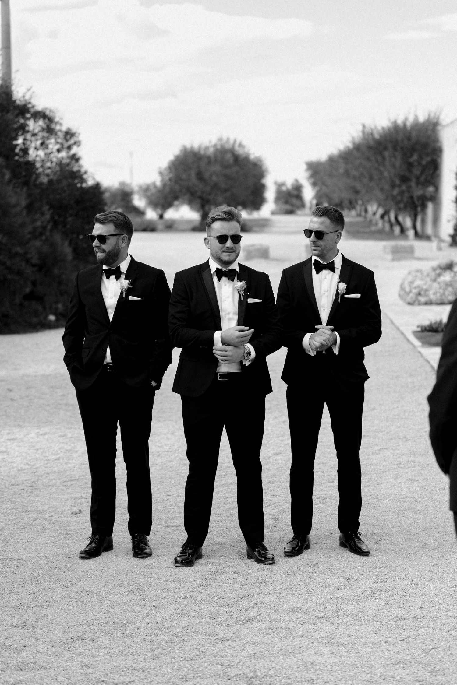 Three groomsmen in classic black tuxedos and sunglasses pose together outdoors.