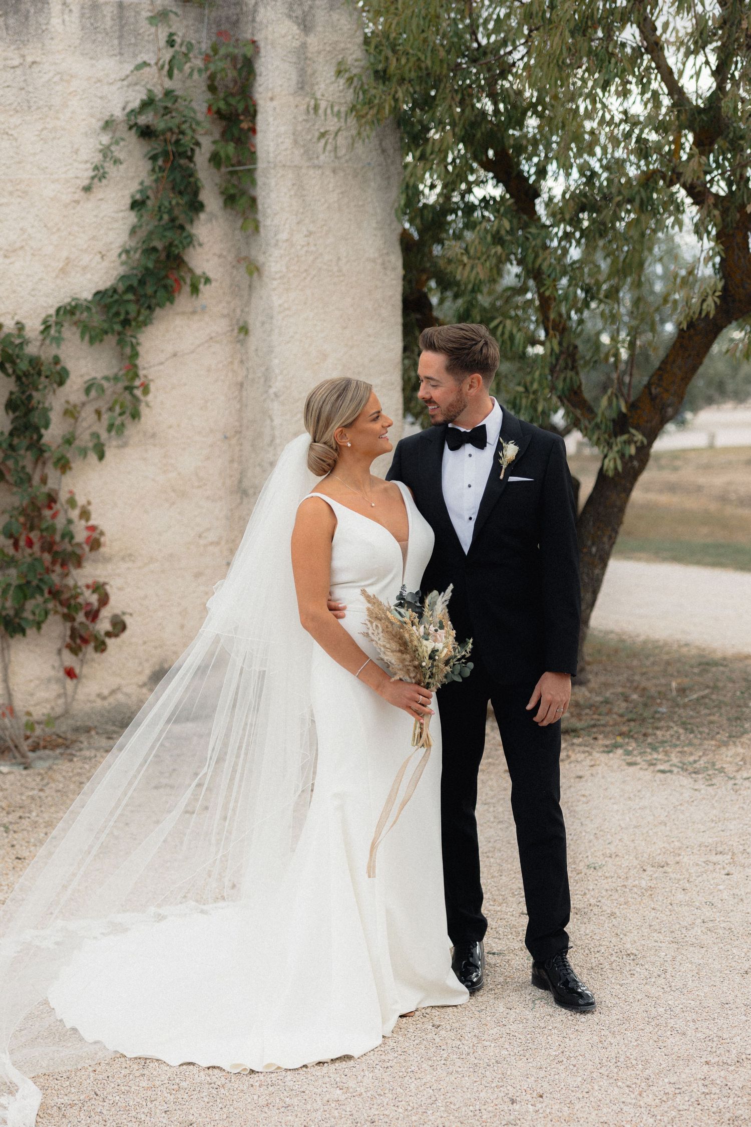 Wedding couple shares intimate moments against a light stone wall with trailing vines.