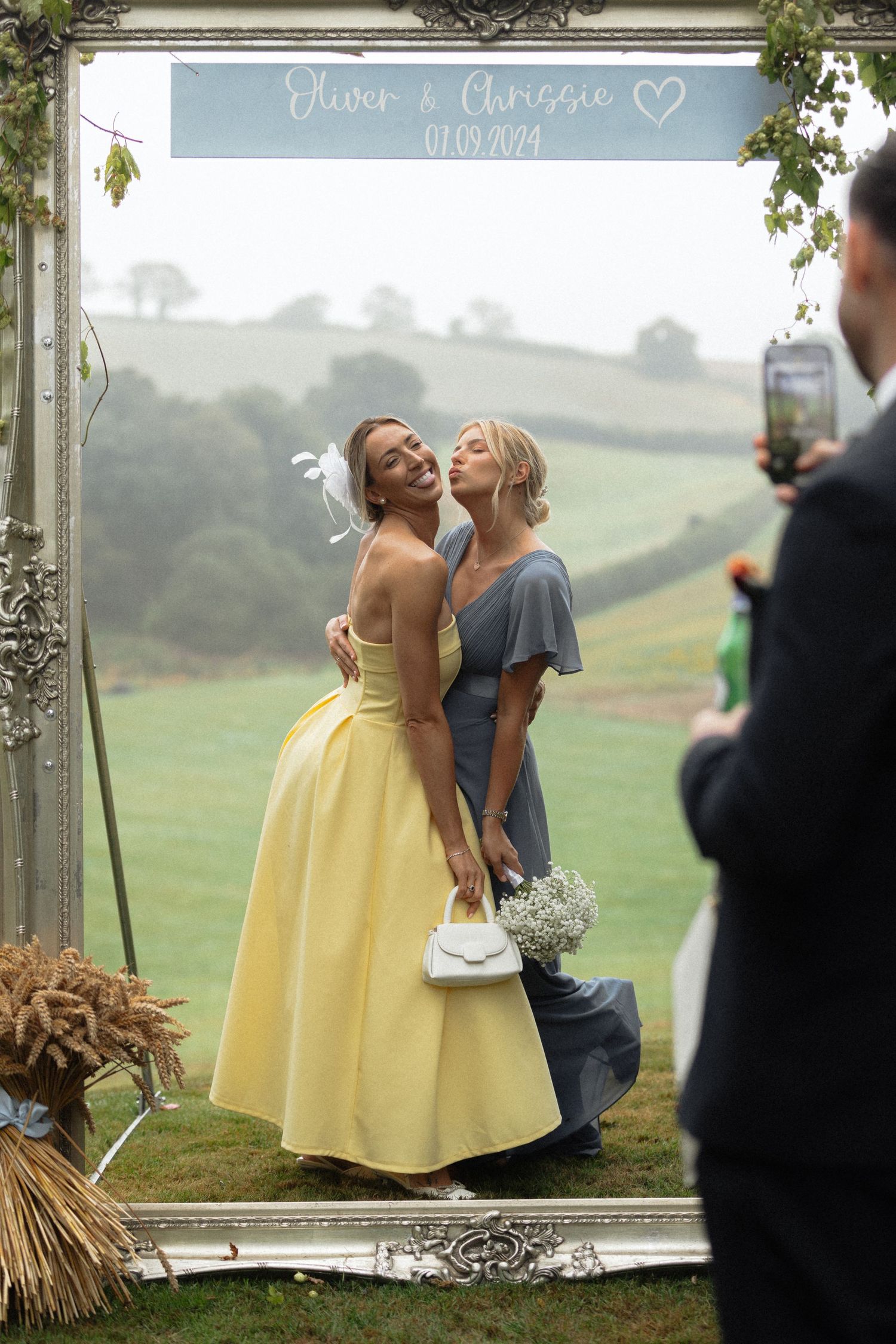 Two people share a joyful moment in front of a mirror at an outdoor wedding venue with rolling hills in the background.