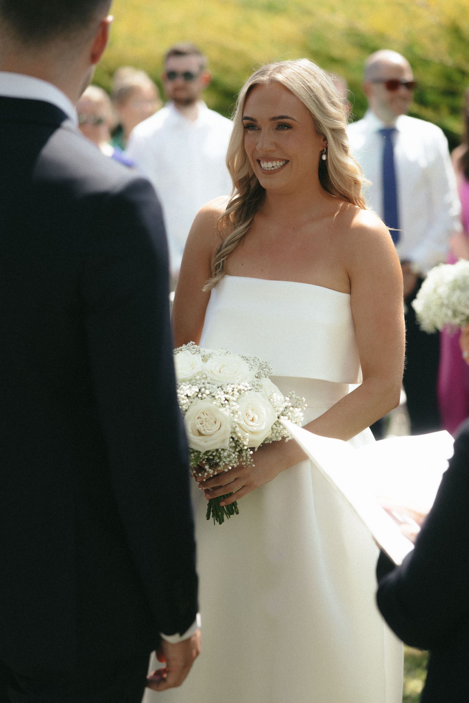Bride in classic white strapless gown holding bouquet during outdoor ceremony.
