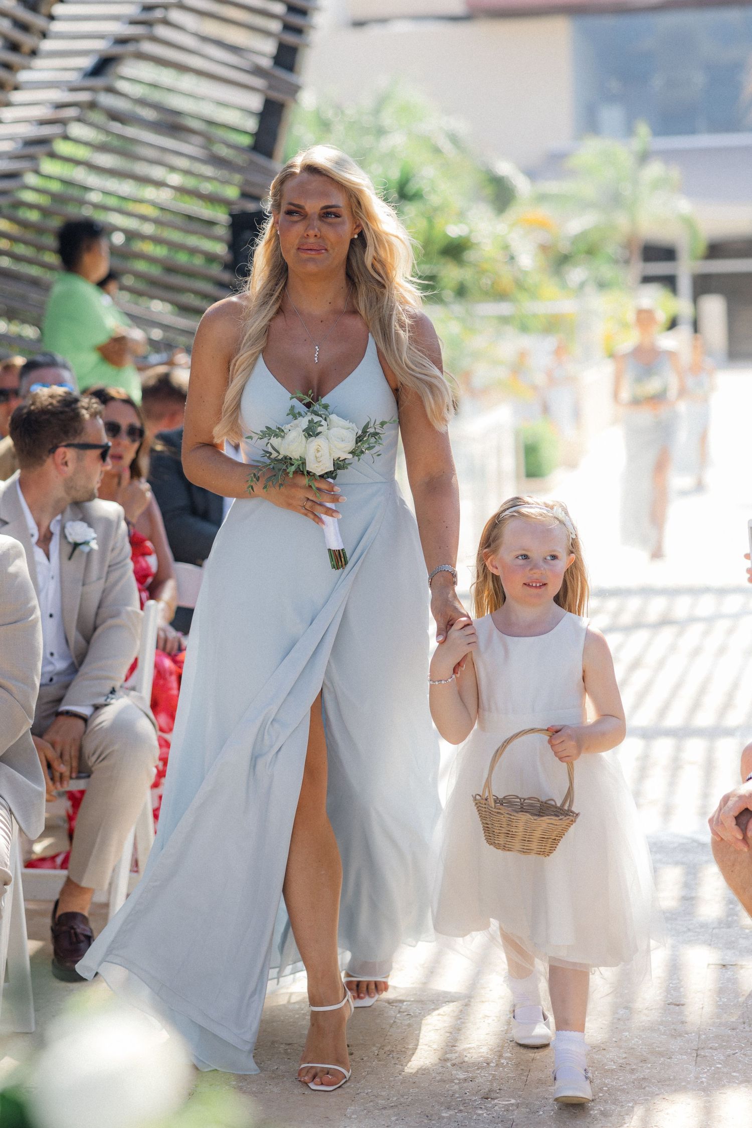 A bride in a light blue dress walks down an outdoor aisle carrying a white bouquet during a sunny wedding ceremony.
