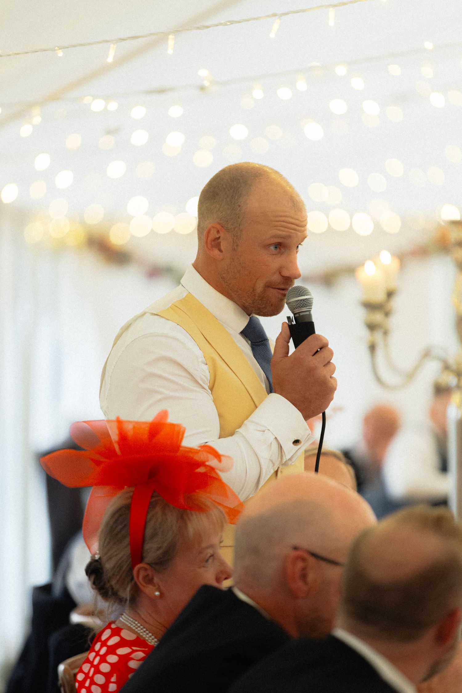 Speaker addresses wedding reception guests under string lights in tent setting.