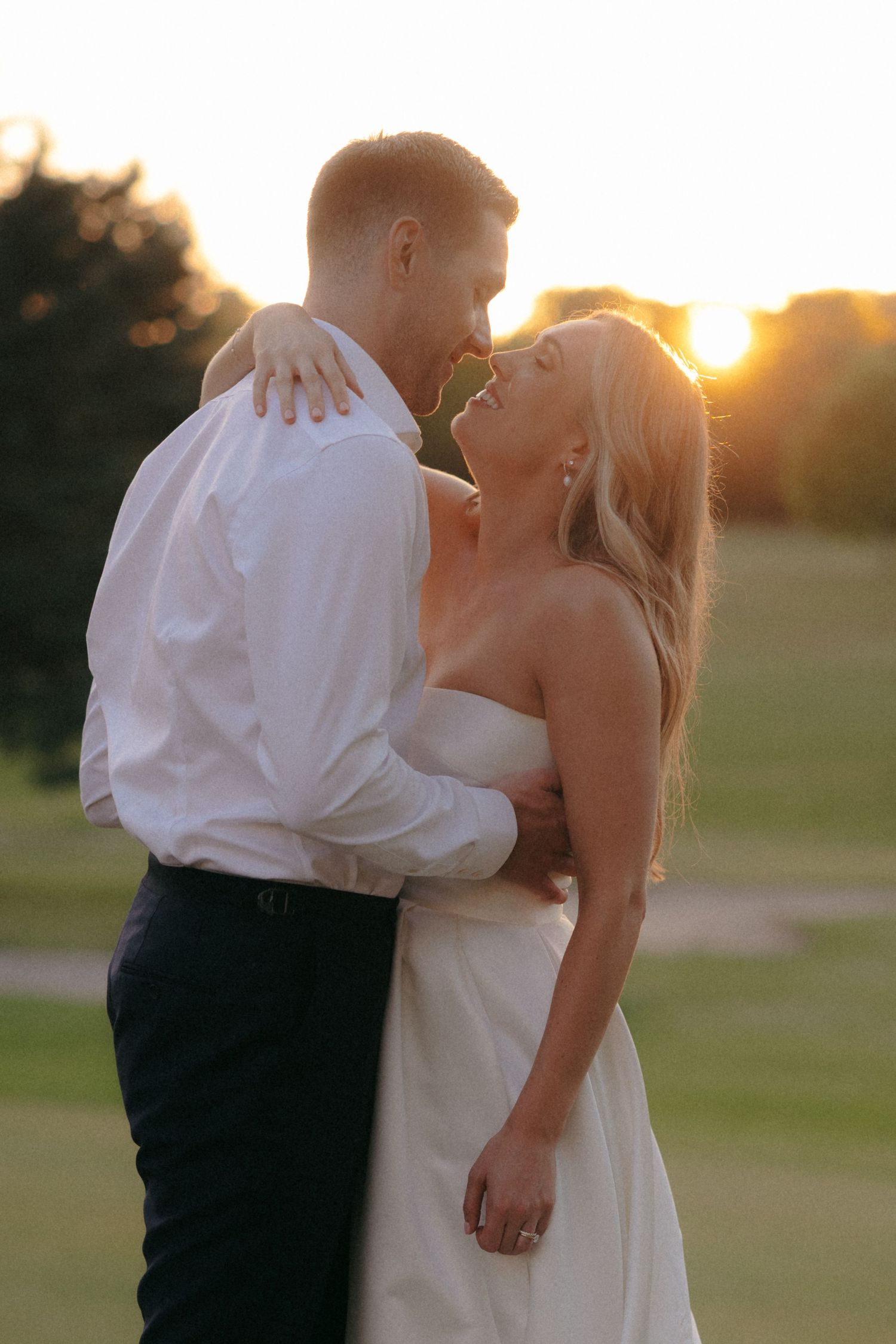 A romantic sunset kiss between newlyweds in formal wedding attire on a grassy field.