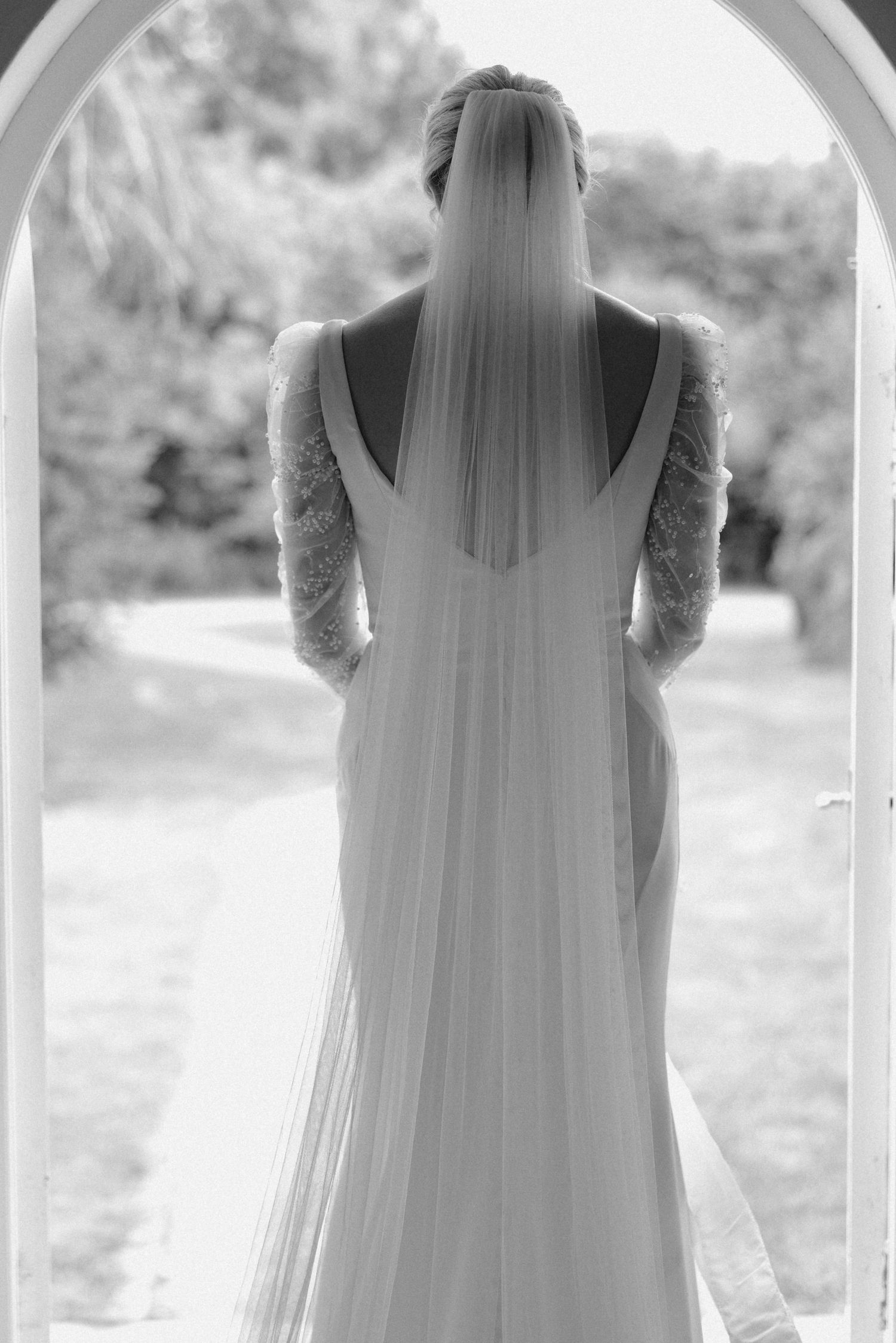 Back view of a bride wearing a flowing veil and lace sleeve wedding gown in a garden setting.