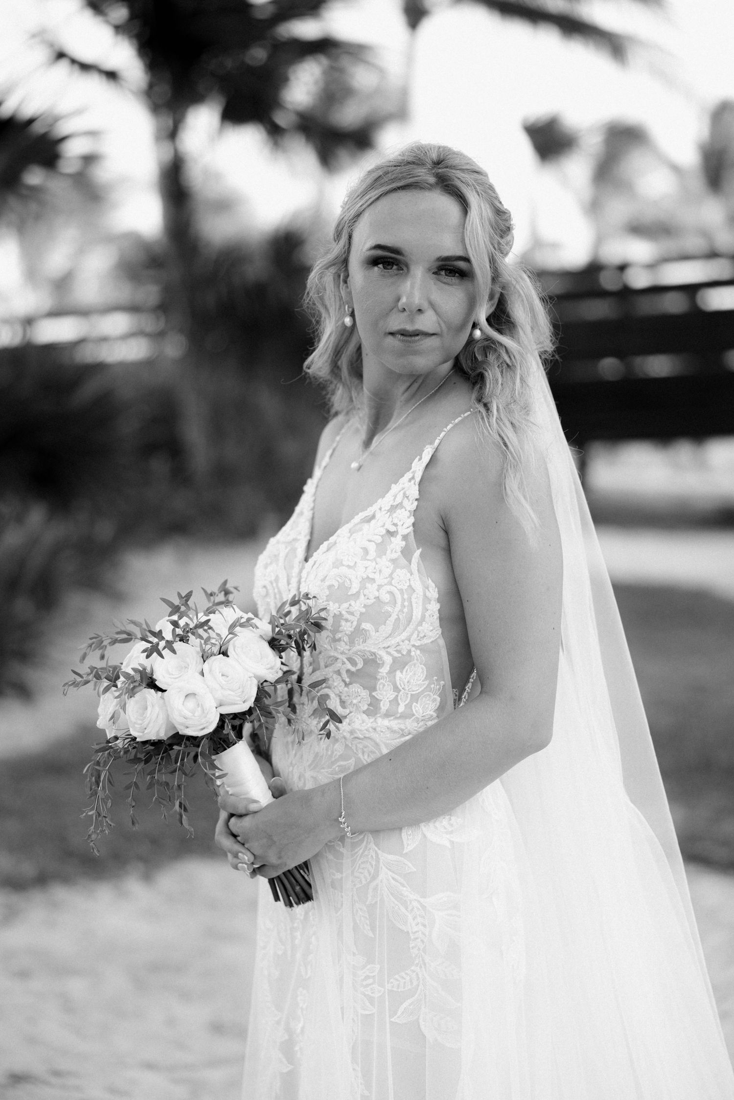 A bride in a white lace dress holds a bouquet of roses in black and white photography.
