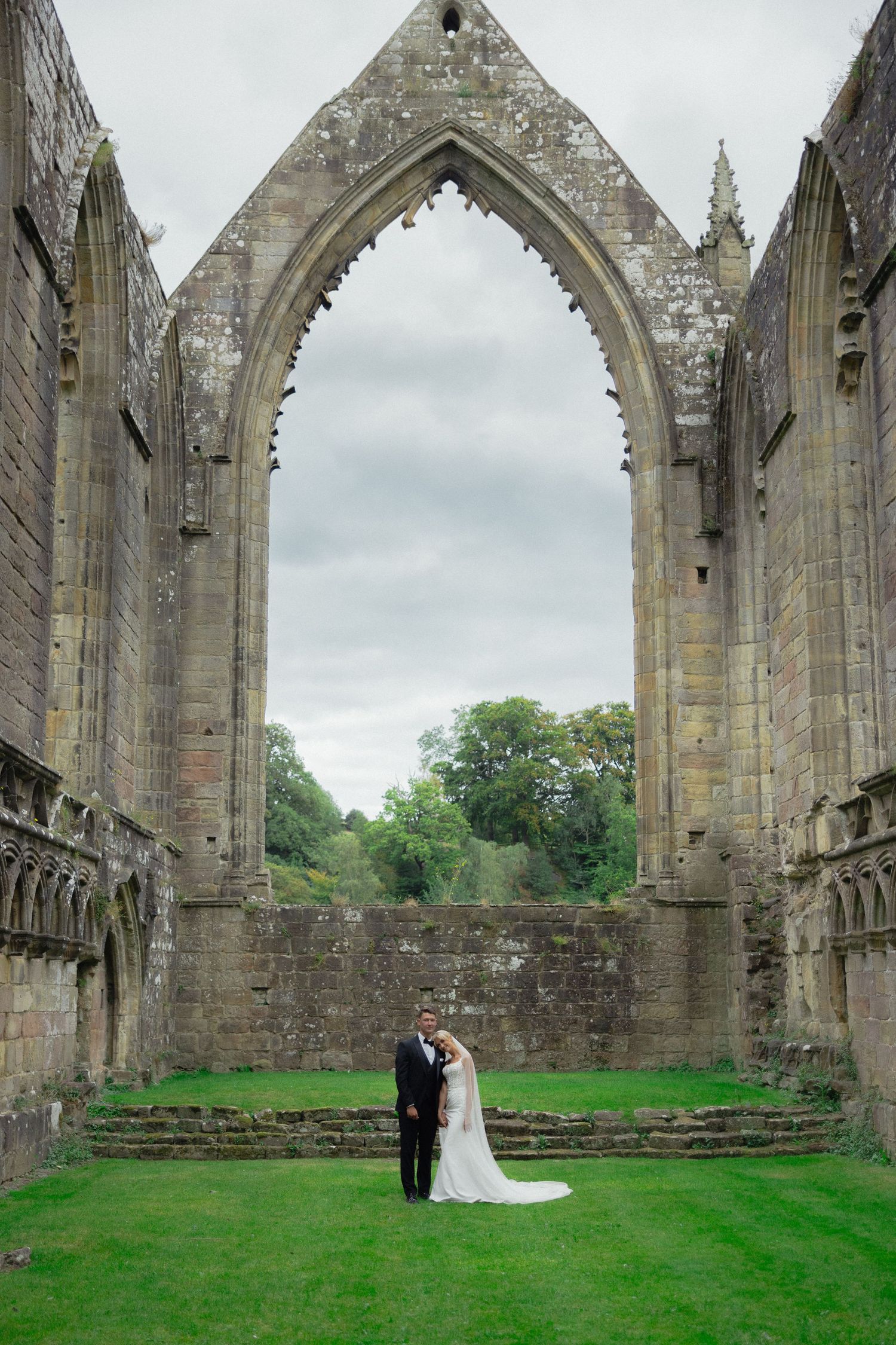 Wedding couple embraces under towering gothic ruins with green lawn.