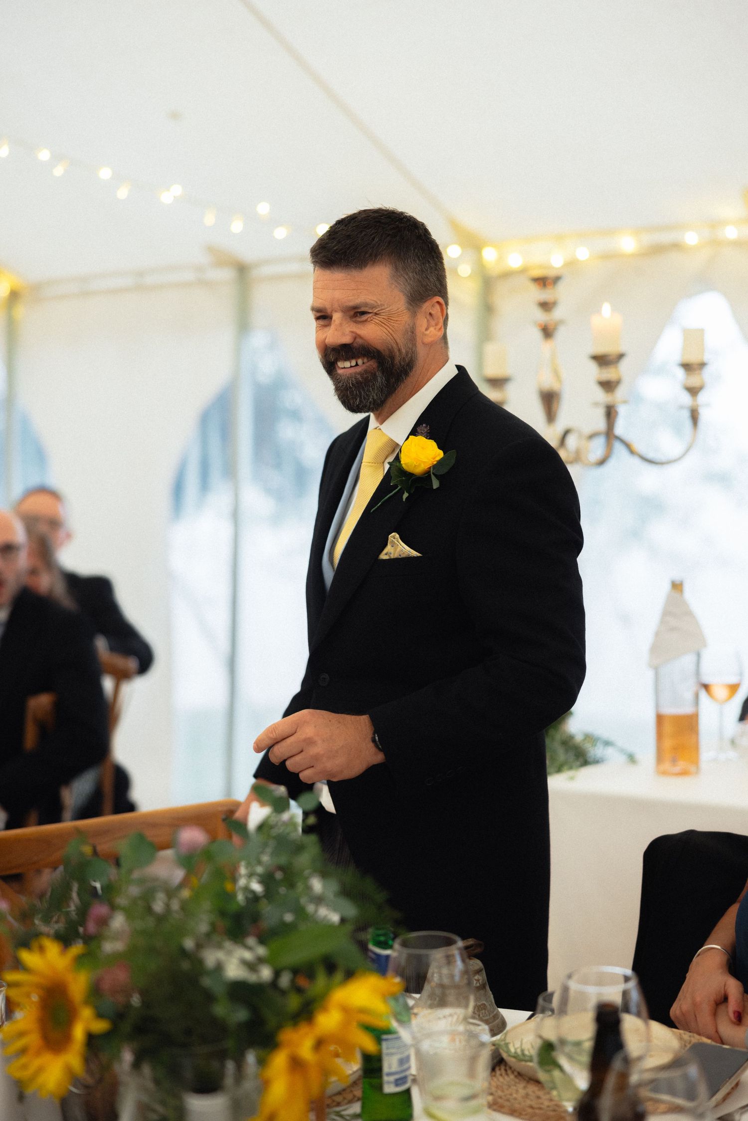 Guest in black suit with yellow boutonniere smiles during wedding reception.
